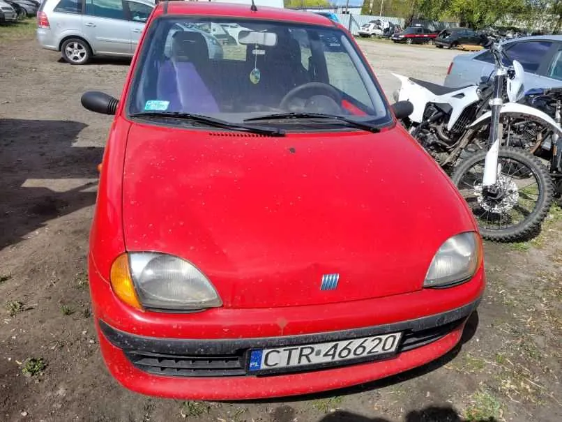 Czerwony samochód osobowy marki Fiat z tablicą rejestracyjną PL CTR-46620, zaparkowany na terenie ziemnym z widocznymi plamami brudu na nadwoziu. Po prawej stronie samochodu stoi biały motocykl enduro. W tle widoczne są inne pojazdy, w tym srebrny minivan i czarne samochody. Na przedniej szybie samochodu znajduje się niebieski znaczek oraz wiszący na lusterku wstecznym ozdobny element. Tło stanowi otwarty teren z ziemią i fragmentami trawy.