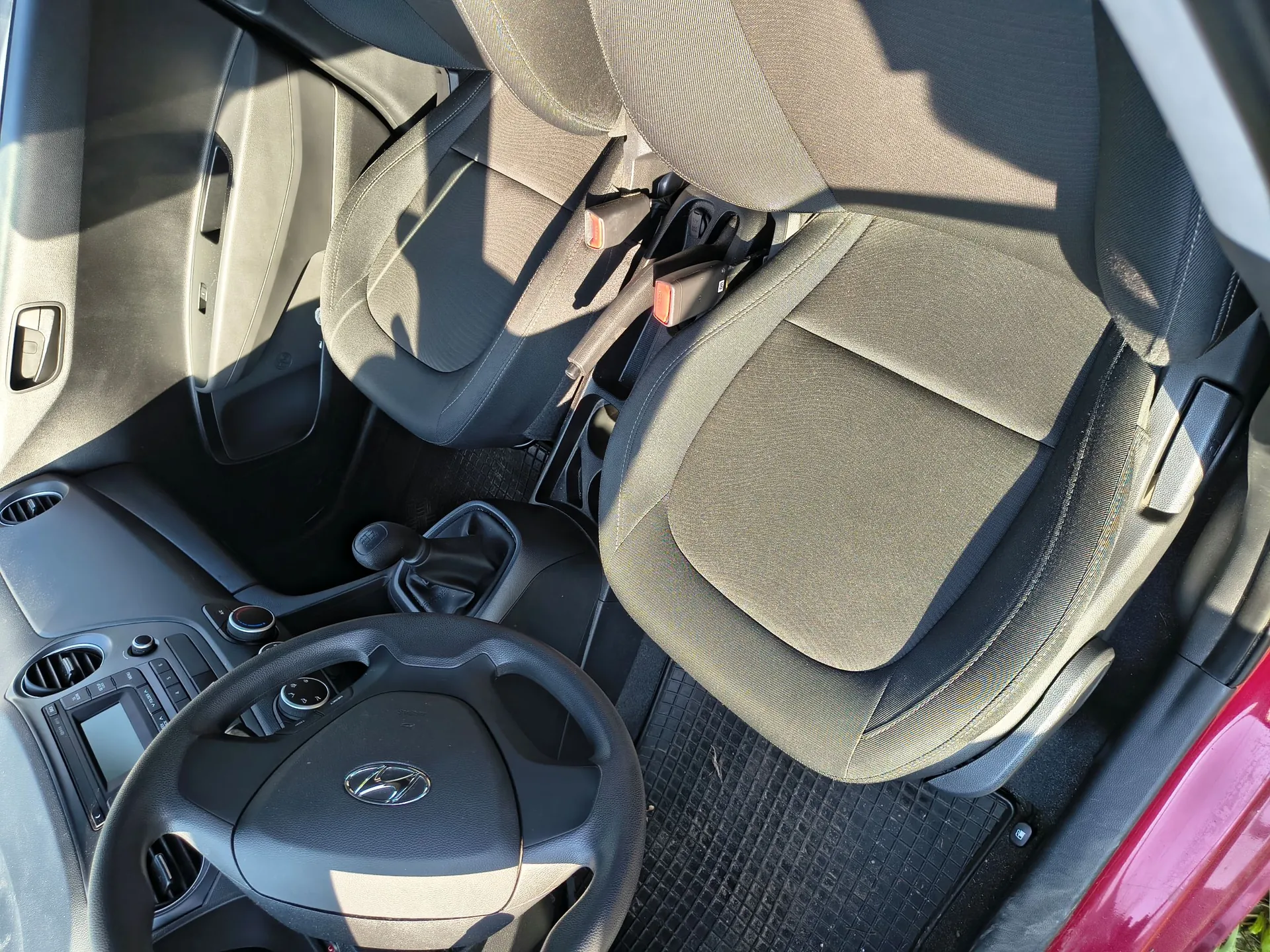 Wnętrze samochodu marki Hyundai z widokiem na przednie fotele z ciemnoszarej tkaniny, deskę rozdzielczą, kierownicę z logo producenta oraz lewarek zmiany biegów. Wnętrze jest oświetlone naturalnym światłem słonecznym.