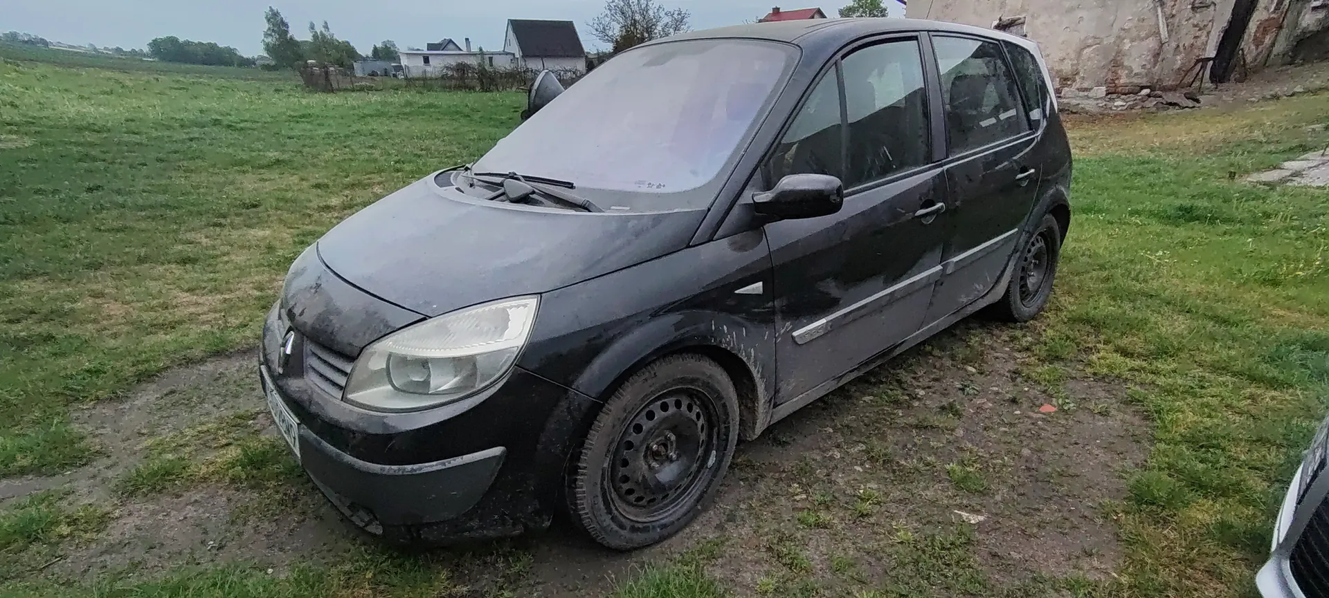 Czarny, zabrudzony samochód osobowy typu minivan zaparkowany na trawiastym i błotnistym terenie. W tle widoczne domy, zielone pole oraz częściowo zniszczona budowla. Samochód ma brudne felgi i zarysowaną karoserię. Na pierwszym planie po prawej stronie widoczna część innego pojazdu.
