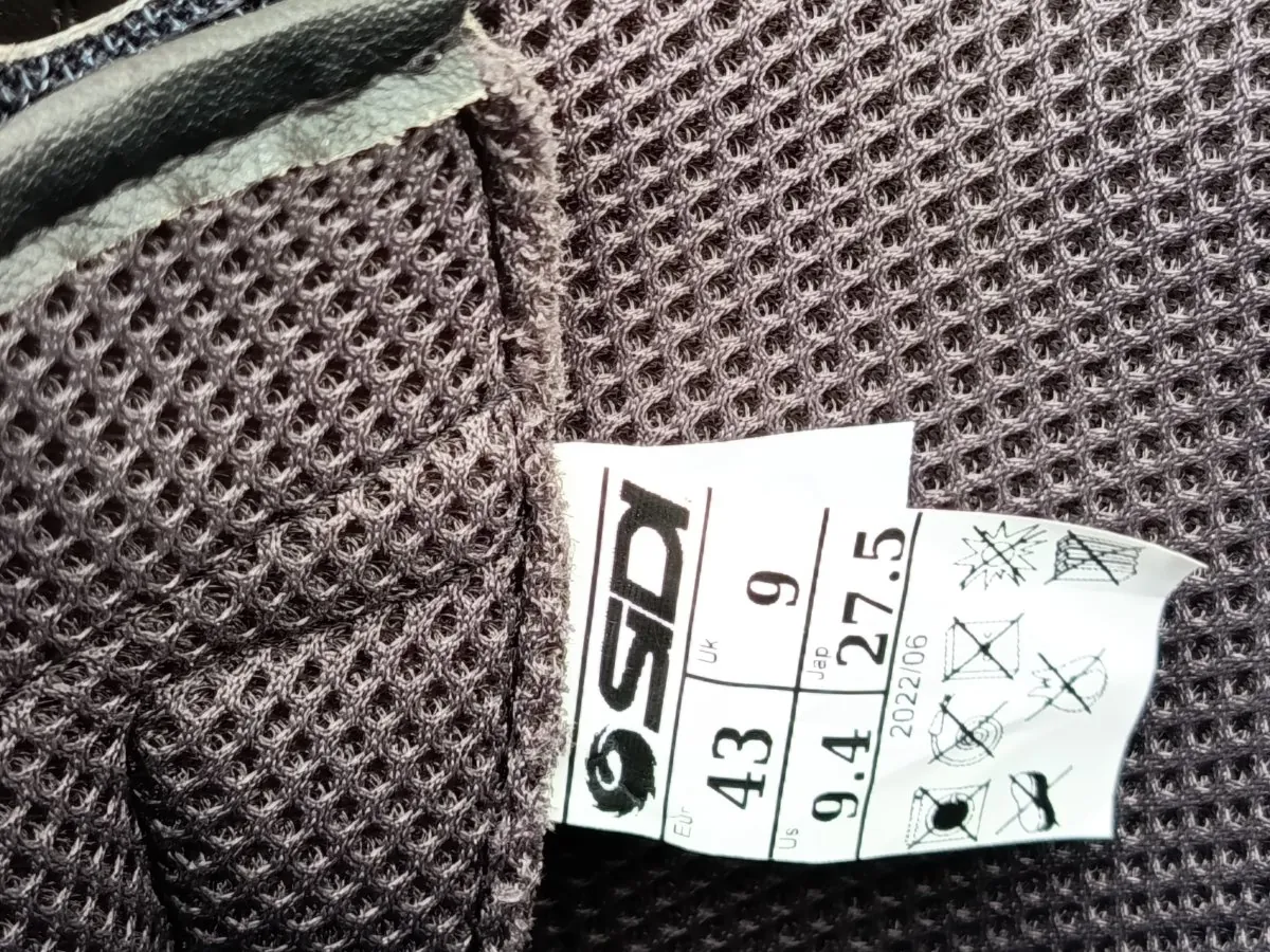 Zbliżenie na wnętrze buta z ciemnoszarej, oddychającej siateczki. Widoczna jest biała metka z logo 'SDI' oraz informacjami o rozmiarach: 'EU 43', 'UK 9', 'US 9.4', 'Jap 27.5'. Na metce znajduje się również data '2022/06' oraz symbole dotyczące konserwacji i pielęgnacji produktu.
