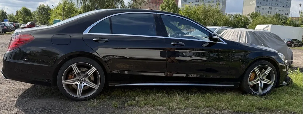 Czarny samochód osobowy marki Mercedes-Benz S-Klasy zaparkowany bokiem na utwardzonym placu z trawą. Pojazd ma srebrne felgi aluminiowe i chromowane wykończenia wokół okien oraz wzdłuż progu. W tle widoczne są inne zaparkowane samochody, z czego jeden jest przykryty szarą plandeką, oraz wielopiętrowe budynki mieszkalne i drzewa.