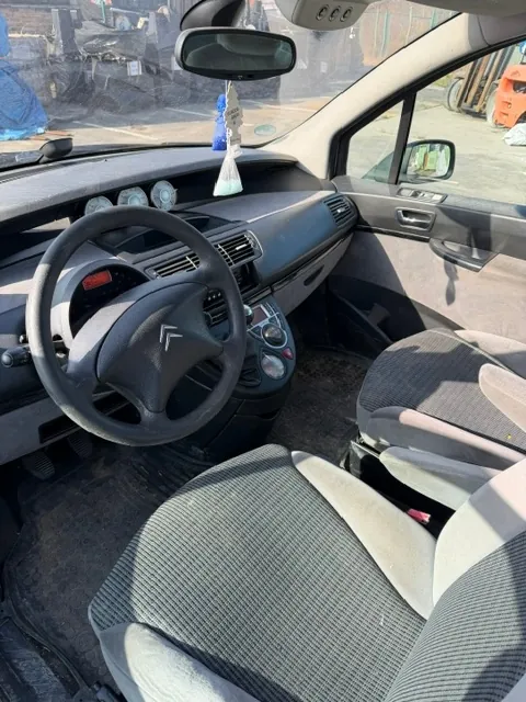 Wnętrze samochodu marki Citroën widziane z perspektywy pasażera. Widoczna jest kierownica z logo marki, deska rozdzielcza, dwa fotele z szaro-czarną tapicerką w prążki oraz lusterko wsteczne, do którego przyczepiony jest niebieski zapach samochodowy.
