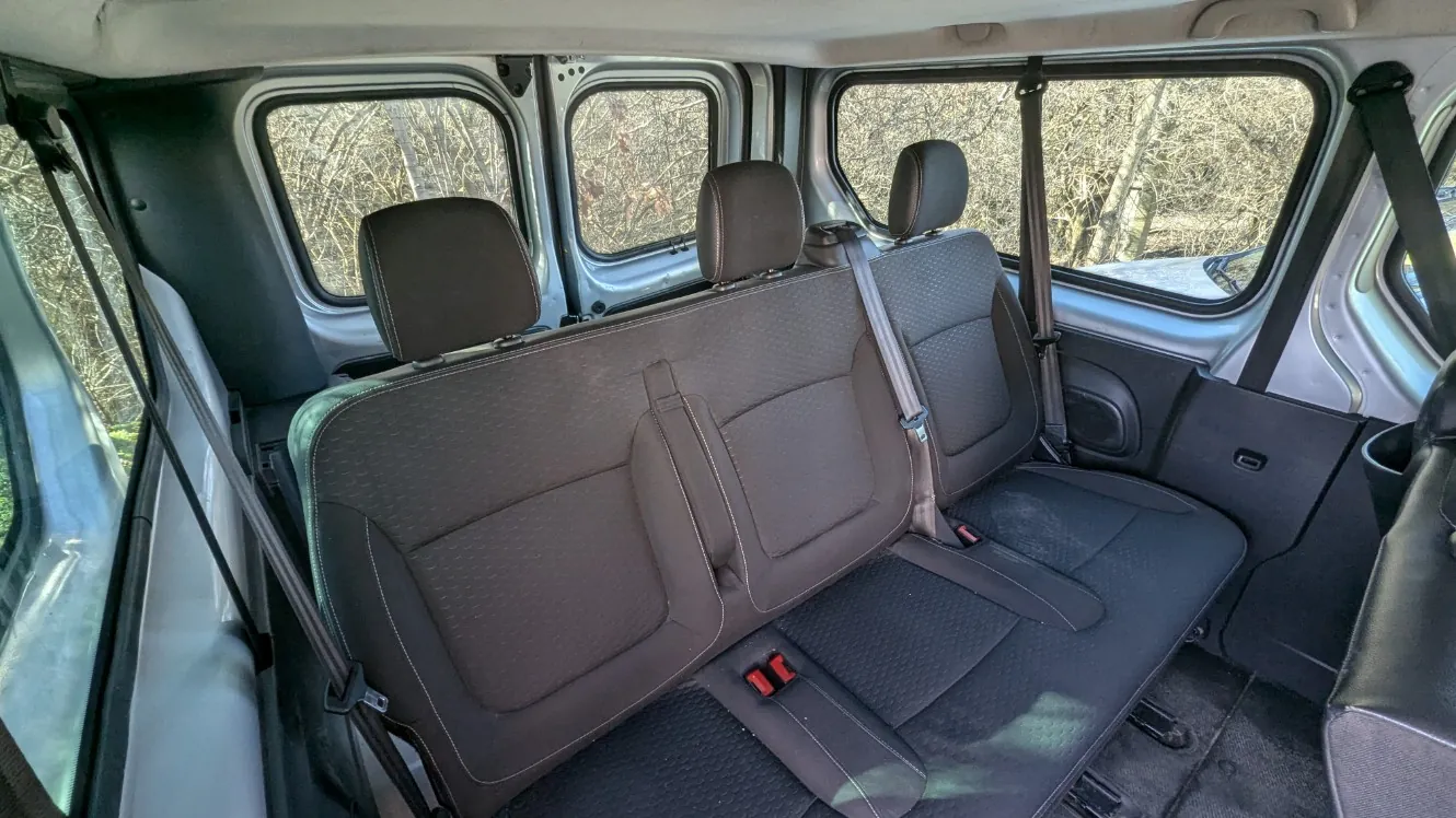 Wnętrze pojazdu widziane od tyłu, przedstawiające trzy siedzenia w drugim rzędzie. Siedzenia są ciemnoszare z wzorzystą tapicerką, wyposażone w pasy bezpieczeństwa z czerwonymi zapięciami. Po bokach widoczne są okna z widokiem na suche drzewa na zewnątrz. Wnętrze posiada szare i ciemnoszare panele, a podłoga jest pokryta ciemnym dywanikiem. Brak czytelnego tekstu na obrazie.