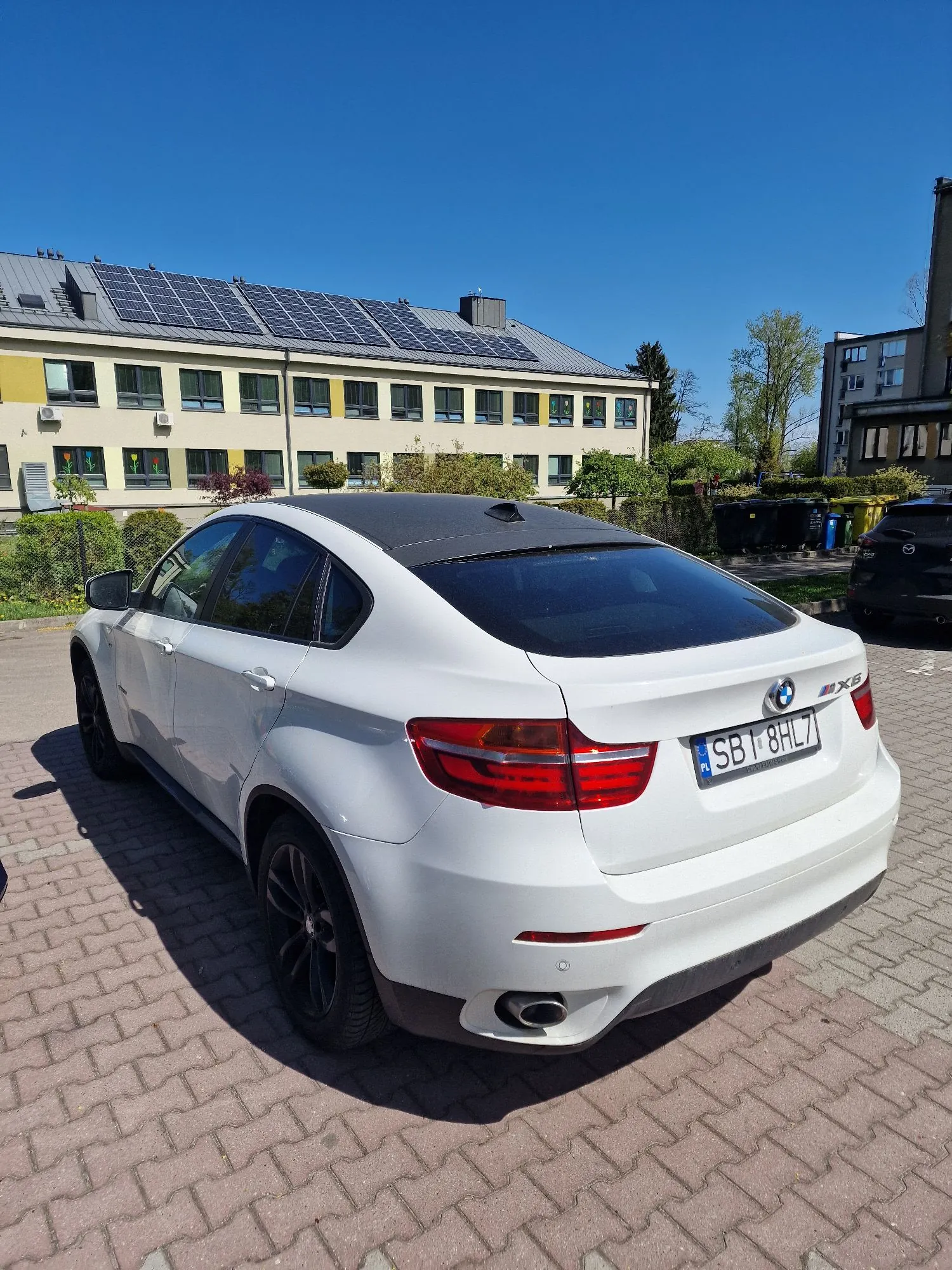 Biały samochód marki BMW X6 zaparkowany tyłem na kostce brukowej. Pojazd ma czarny dach i czarne felgi. Tablica rejestracyjna to SBI 8HL7. W tle widoczny jest dwupiętrowy żółty budynek z panelami fotowoltaicznymi na dachu, zielone krzewy oraz bezchmurne, niebieskie niebo.