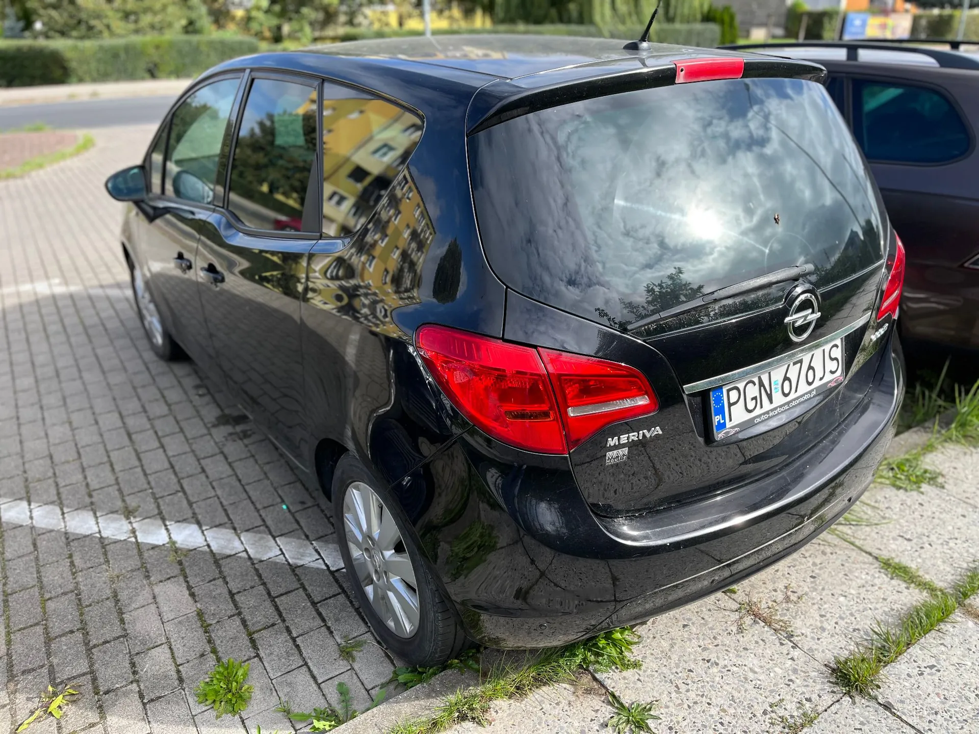 Czarny samochód marki Opel Meriva zaparkowany tyłem na kostce brukowej. Widoczna jest tylna część pojazdu z tablicą rejestracyjną PGN 676JS oraz logo producenta. W tle widać fragmenty zieleni i budynków odbijające się w karoserii i szybach auta.