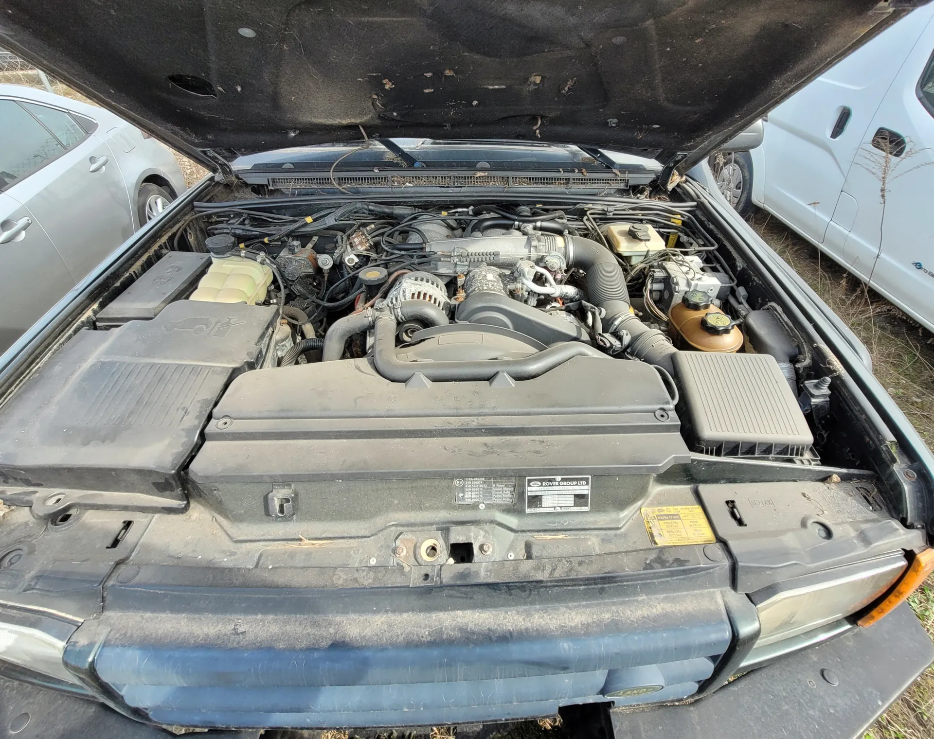 Samochód z otwartą maską ukazujący komorę silnika z różnymi elementami: przewodami, zbiornikami (w tym żółtym i brązowym), czarnymi osłonami oraz tabliczkami z napisami. Widać tabliczkę z napisem 'ROVER GROUP LTD' i żółtą naklejkę z nieczytelnym tekstem. W tle stoją dwa inne pojazdy – srebrny i biały samochód. Scena rozgrywa się na zewnątrz, na trawie i suchym terenie.