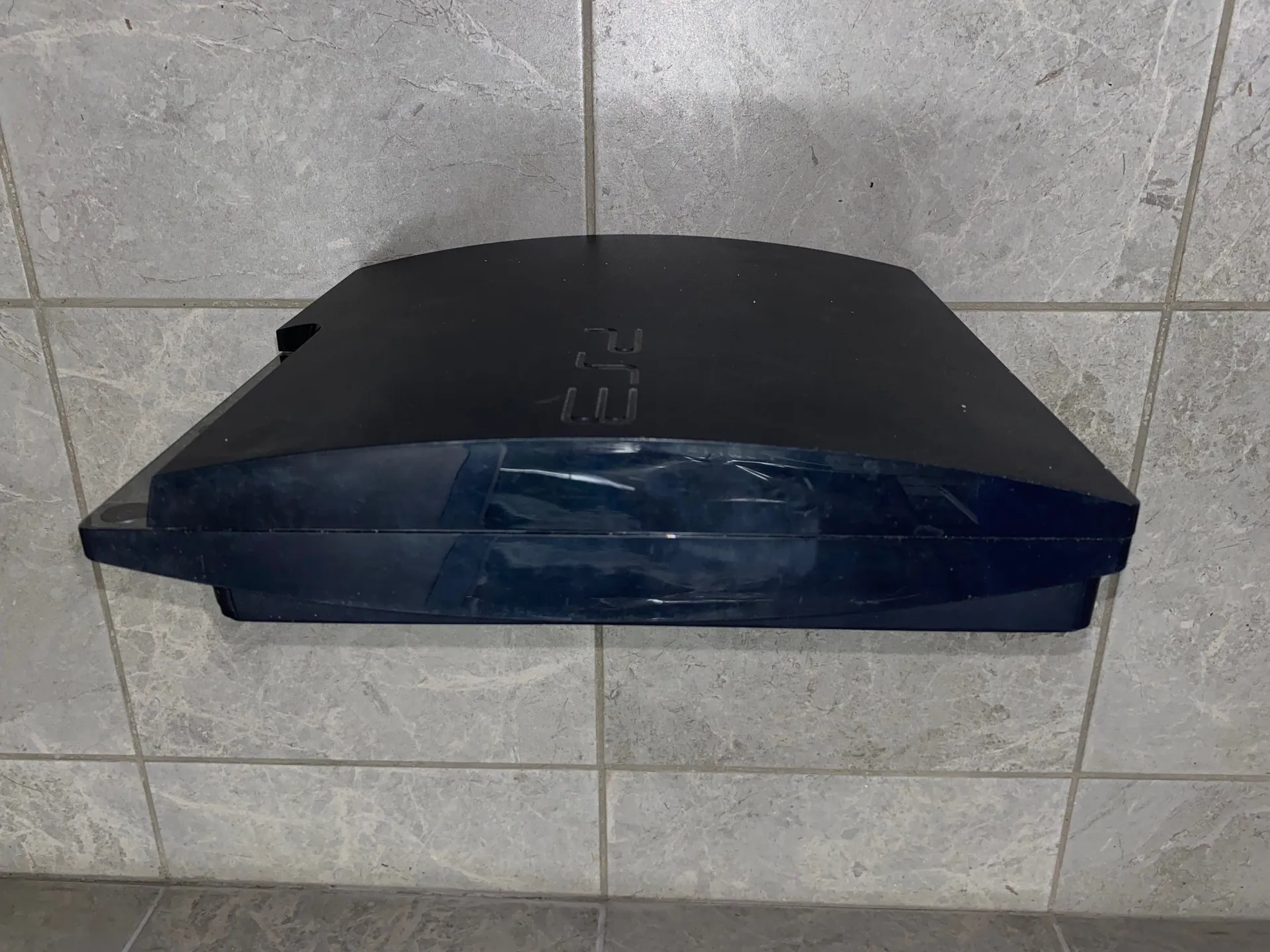 Czarny, plastikowy dozownik do papierowych ręczników zamontowany na szarej, kafelkowej ścianie. Na górnej części urządzenia znajduje się wytłoczony napis 'Tork'.