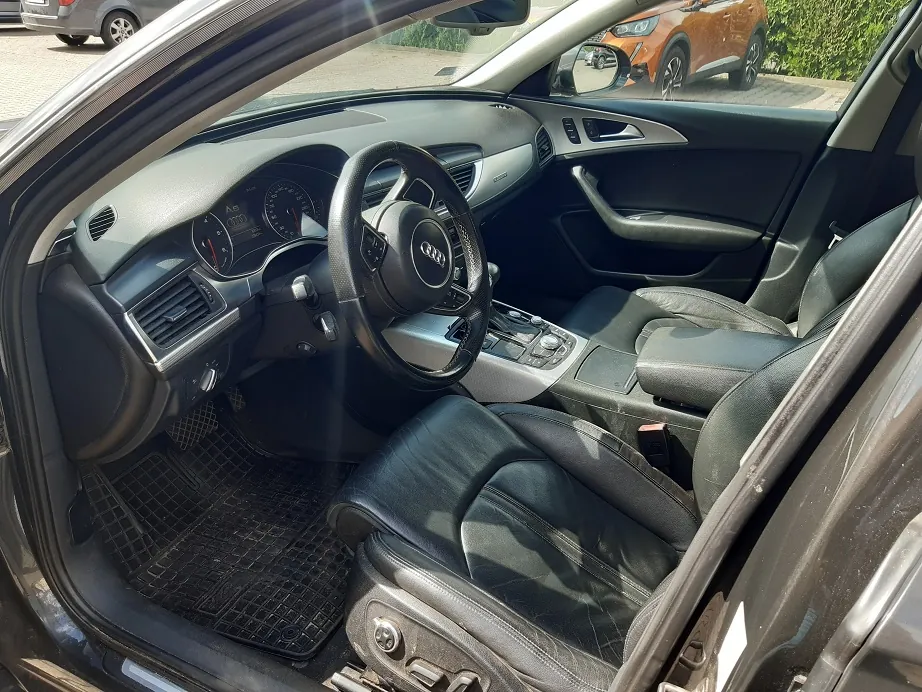 Wnętrze samochodu marki Audi widziane z otwartych drzwi kierowcy. Widoczna jest czarna skórzana tapicerka foteli, czarna kierownica z logo Audi, deska rozdzielcza z zegarami oraz konsola środkowa. W tle, przez szybę, widoczny jest fragment pomarańczowego samochodu zaparkowanego na zewnątrz.