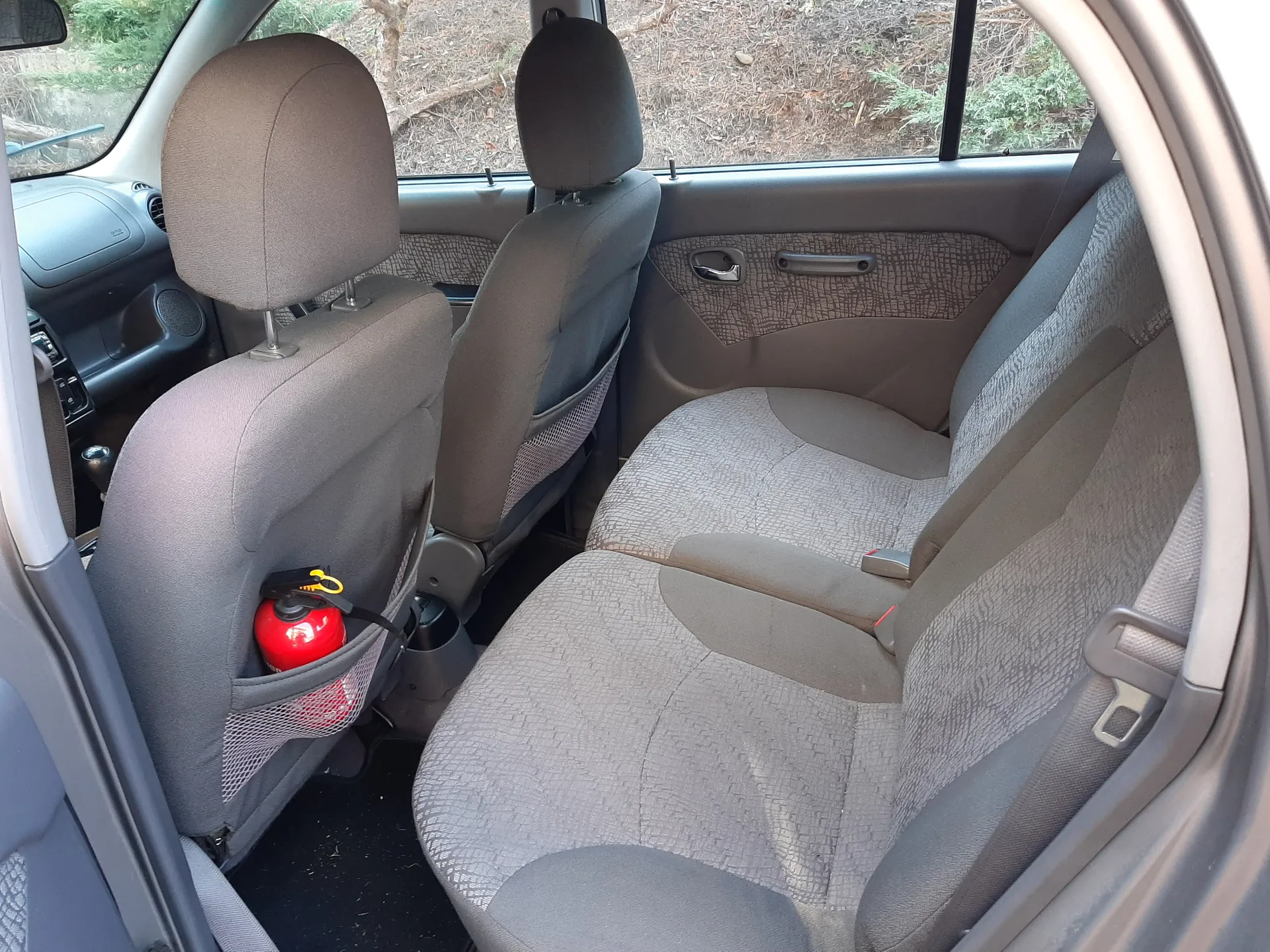 Wnętrze samochodu widziane od tyłu. Widoczne są dwa przednie siedzenia w kolorze szaro-brązowym z wzorzystą tapicerką. W siateczce na oparciu siedzenia pasażera znajduje się czerwony gaśnicz z żółtym elementem. Drzwi po stronie kierowcy są otwarte, a przez okna widać zewnętrzny krajobraz z suchą ziemią i roślinnością. W tle widoczne są fragmenty deski rozdzielczej oraz uchwytów drzwiowych.