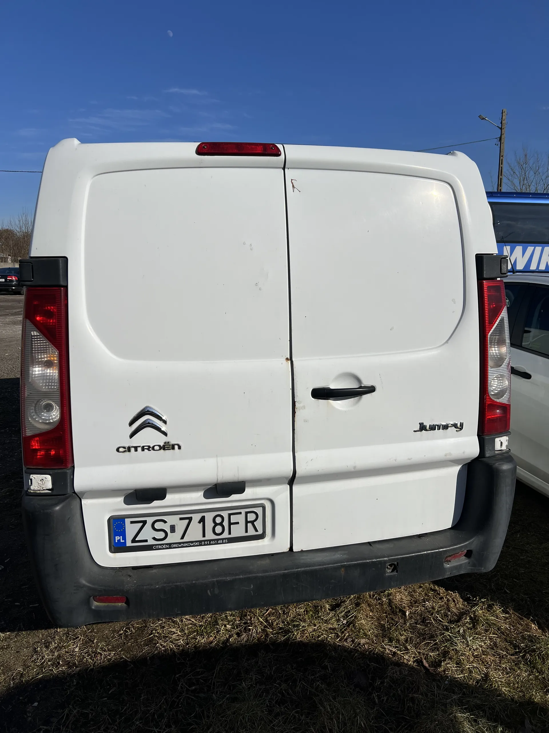 Tył białego furgonetki Citroën Jumpy z tablicą rejestracyjną ZS 718FR. Widoczne są czerwone światła tylne, logo Citroën oraz czarny zderzak. W tle niebieskie niebo i trawa. Częściowo widoczny inny pojazd z niebieskim napisem WIN.