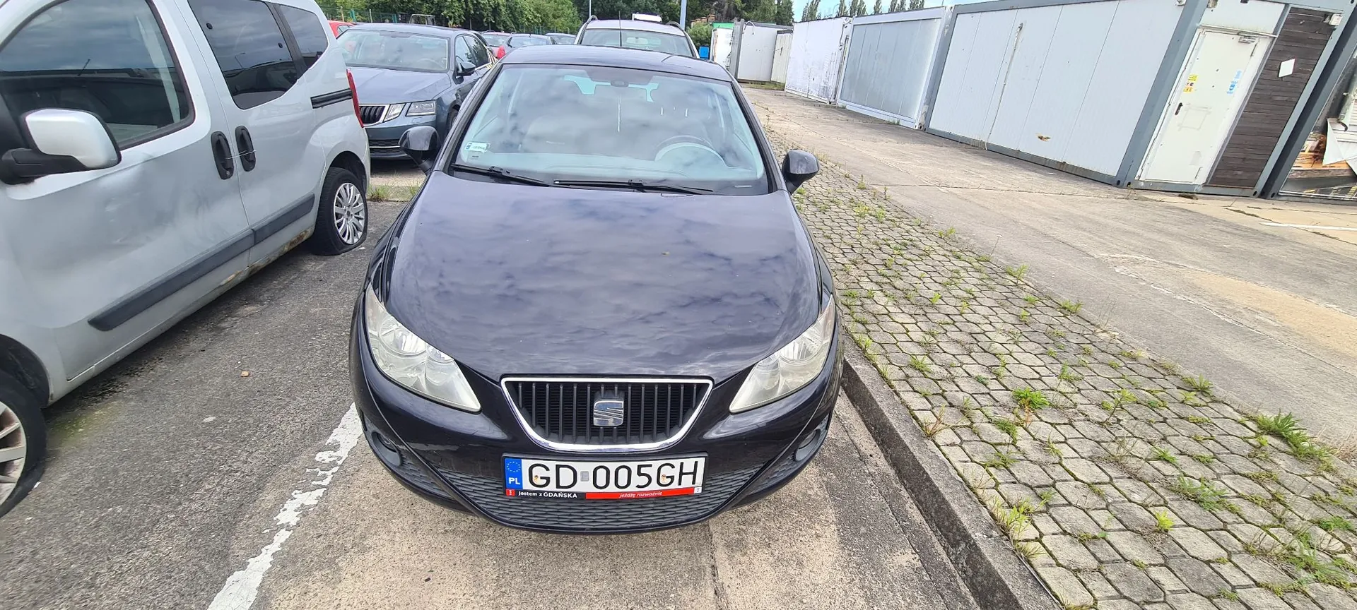 Czarny samochód osobowy marki Seat zaparkowany przodem do obiektywu na betonowym placu. Samochód ma tablicę rejestracyjną o numerze GD 005GH. Po lewej stronie widoczny jest fragment białego vana, a w tle inny ciemny samochód oraz białe kontenery magazynowe.