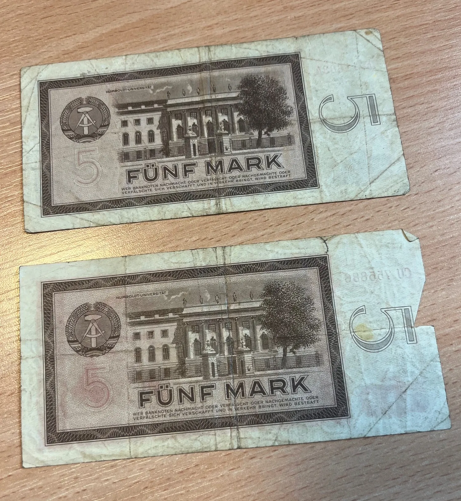 Dwa stare banknoty o nominale 5 marek (Fünf Mark), ułożone jeden nad drugim na drewnianym blacie. Na banknotach widnieje rysunek budynku Uniwersytetu Humboldta (Humboldt-Universität) oraz duża cyfra 5. Na dole znajduje się napis: 'WER BANKNOTEN NACHMACHT ODER VERFÄLSCHT ODER NACHGEMACHTE ODER VERFÄLSCHTE SICH VERSCHAFFT UND IN VERKEHR BRINGT, WIRD BESTRAFT'. Dolny banknot jest częściowo naderwany po prawej stronie.