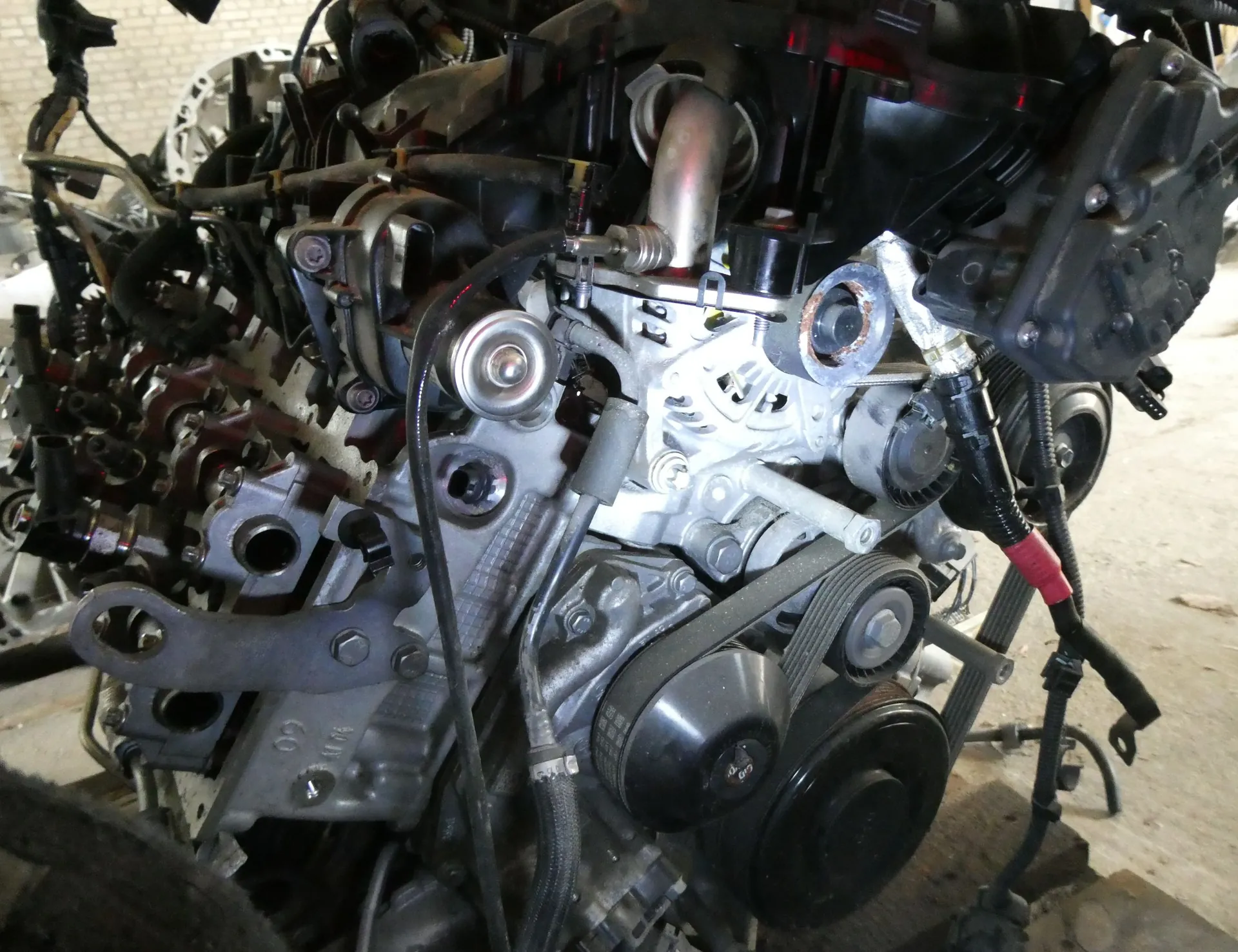 Zbliżenie na złożony silnik spalinowy z widocznymi elementami mechanicznymi, takimi jak paski napędowe, koła pasowe, przewody, rurki i metalowe odlewy bloku silnika. Części są w kolorach srebrnym, czarnym i ciemnoszarym.