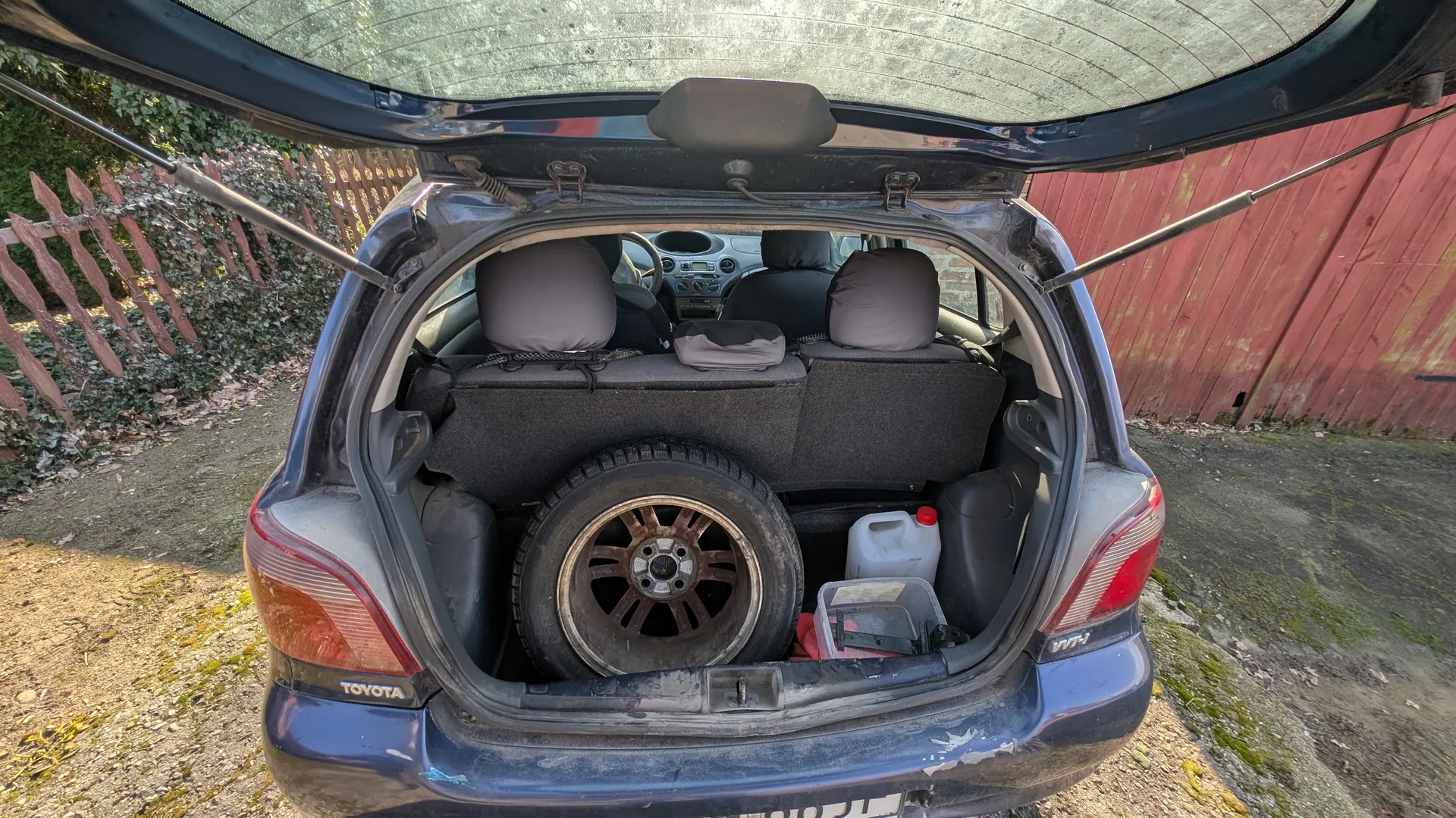 Otwarty bagażnik niebieskiego samochodu marki Toyota. W bagażniku znajduje się zapasowe koło, biały kanister z czerwoną pokrywką oraz przezroczysty plastikowy pojemnik z przedmiotami. Widoczne są czarne siedzenia, kierownica i deska rozdzielcza. Na zewnątrz, w tle, czerwone drewniane ogrodzenie, zielone pnącza i ziemia z suchymi liśćmi. Na tylnej części samochodu widnieje napis "TOYOTA".