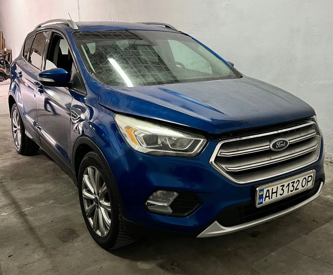 Niebieski samochód typu SUV marki Ford zaparkowany w pomieszczeniu z betonową podłogą i szarą ścianą. Widoczna tablica rejestracyjna z napisem AH 3132 OP oraz flagą Ukrainy po lewej stronie. Na masce umieszczony jest logo Ford. Samochód posiada dachowe pręty i aluminiowe felgi. W tle częściowo widoczny inny pojazd.