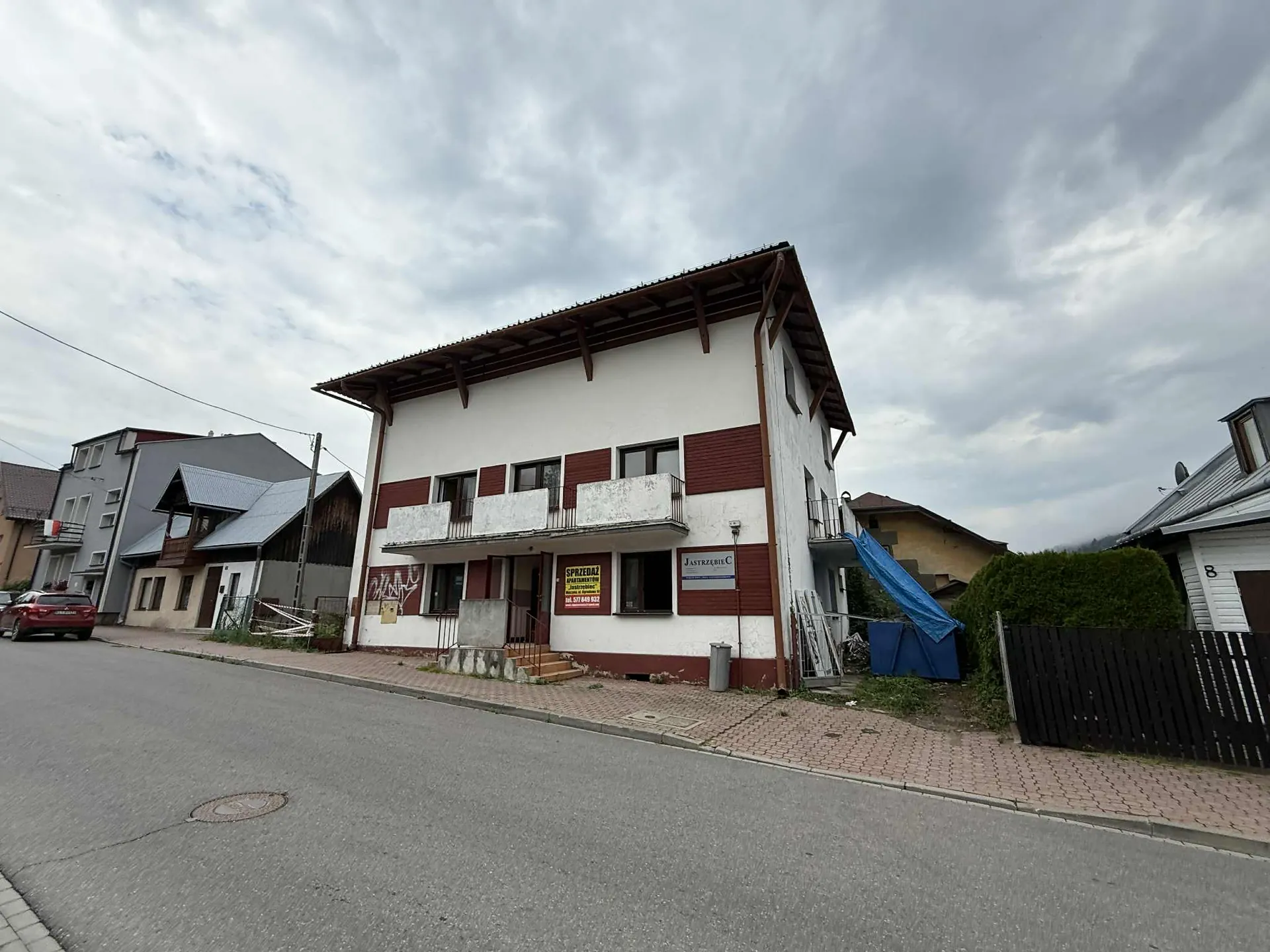 Budynek dwupiętrowy z białymi i brązowymi panelami, z balkonami na pierwszym piętrze. Na ścianie widoczne tablice: żółta z napisem 'SPRZEDAŻ' oraz biała z napisem 'Jastrzębie C'. Przy budynku stoją schody, pojemnik na śmieci, niebieska folia i czarny płot. Po lewej stronie czerwony samochód zaparkowany przy drodze, w tle inne domy i chmurne niebo. Na ścianie budynku widoczne graffiti.