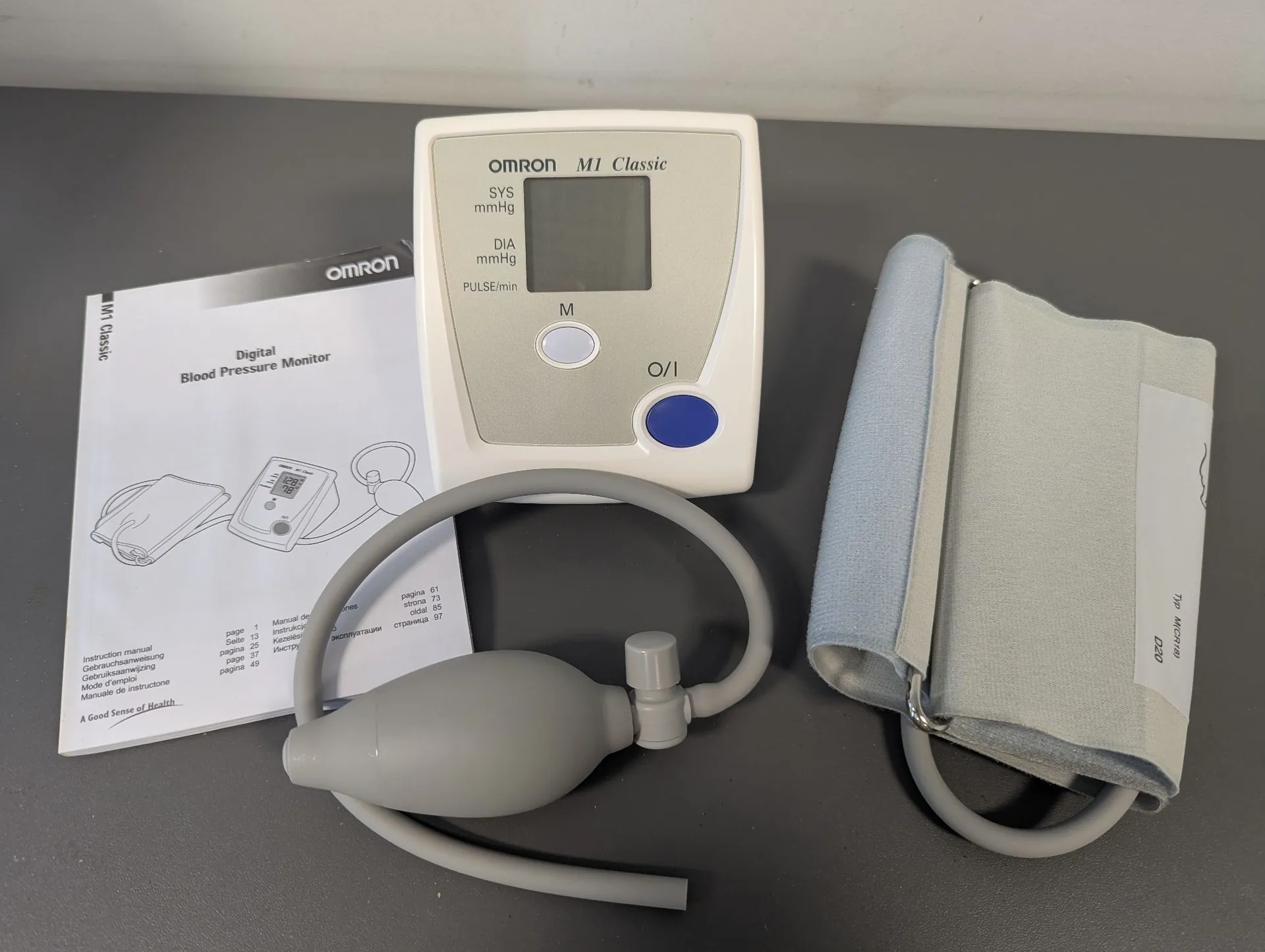 Zestaw ciśnieniomierza elektronicznego marki Omron M1 Classic ułożony na szarym blacie. W skład zestawu wchodzi białe urządzenie z wyświetlaczem LCD i niebieskim przyciskiem zasilania, szary mankiet do ramienia oraz instrukcja obsługi z napisem 'Digital Blood Pressure Monitor'. Urządzenie jest połączone z mankietem za pomocą szarego wężyka z pompką.