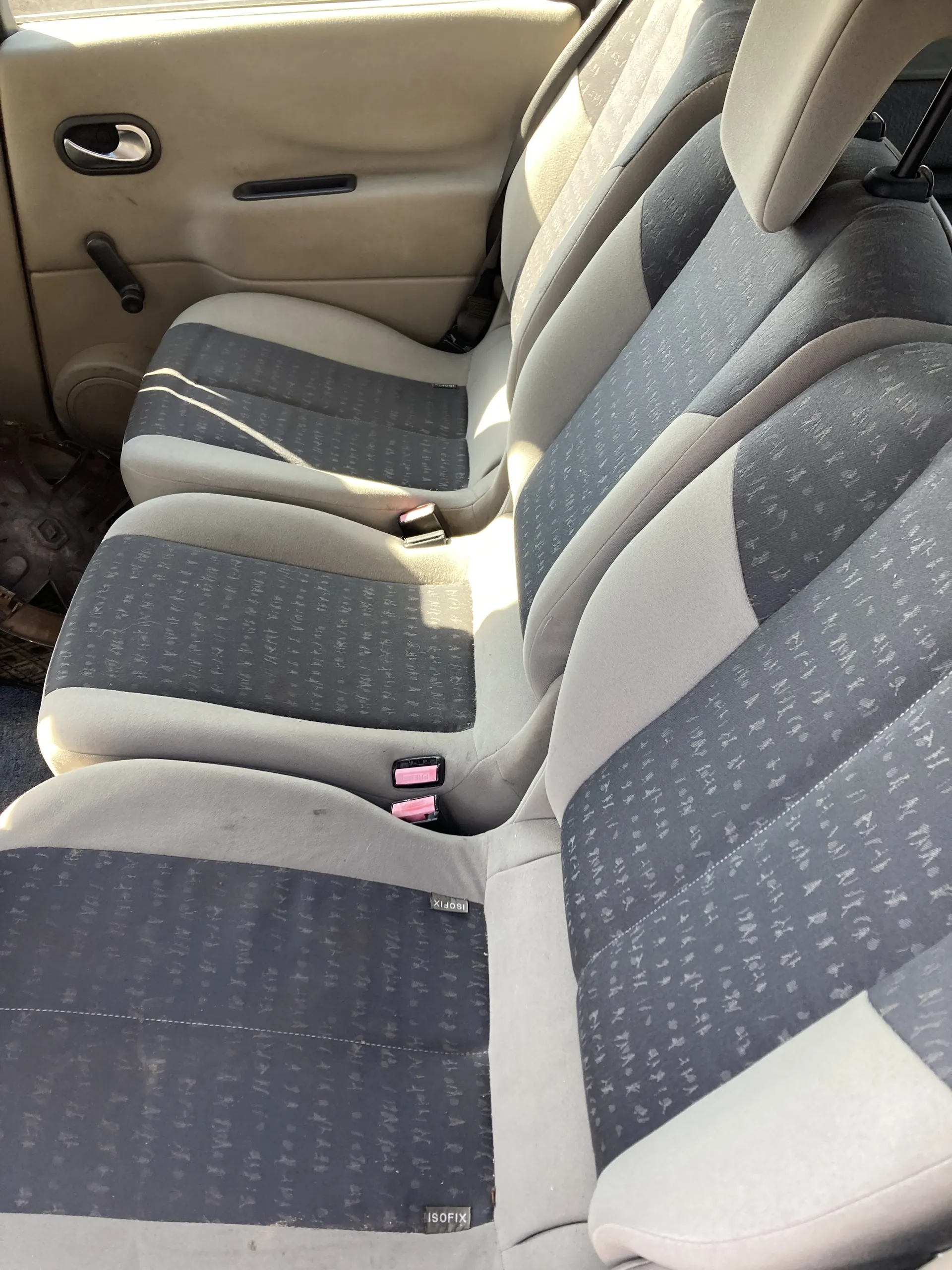 Wnętrze samochodu z widokiem na tylną kanapę. Siedzenia pokryte szarym materiałem z powtarzającym się wzorem, boczne panele w kolorze beżowym. Widoczne różowe zapięcia pasów bezpieczeństwa oraz etykiety ISOFIX na siedzeniach. Drzwi z uchwytem i sterowaniem szyby po lewej stronie.