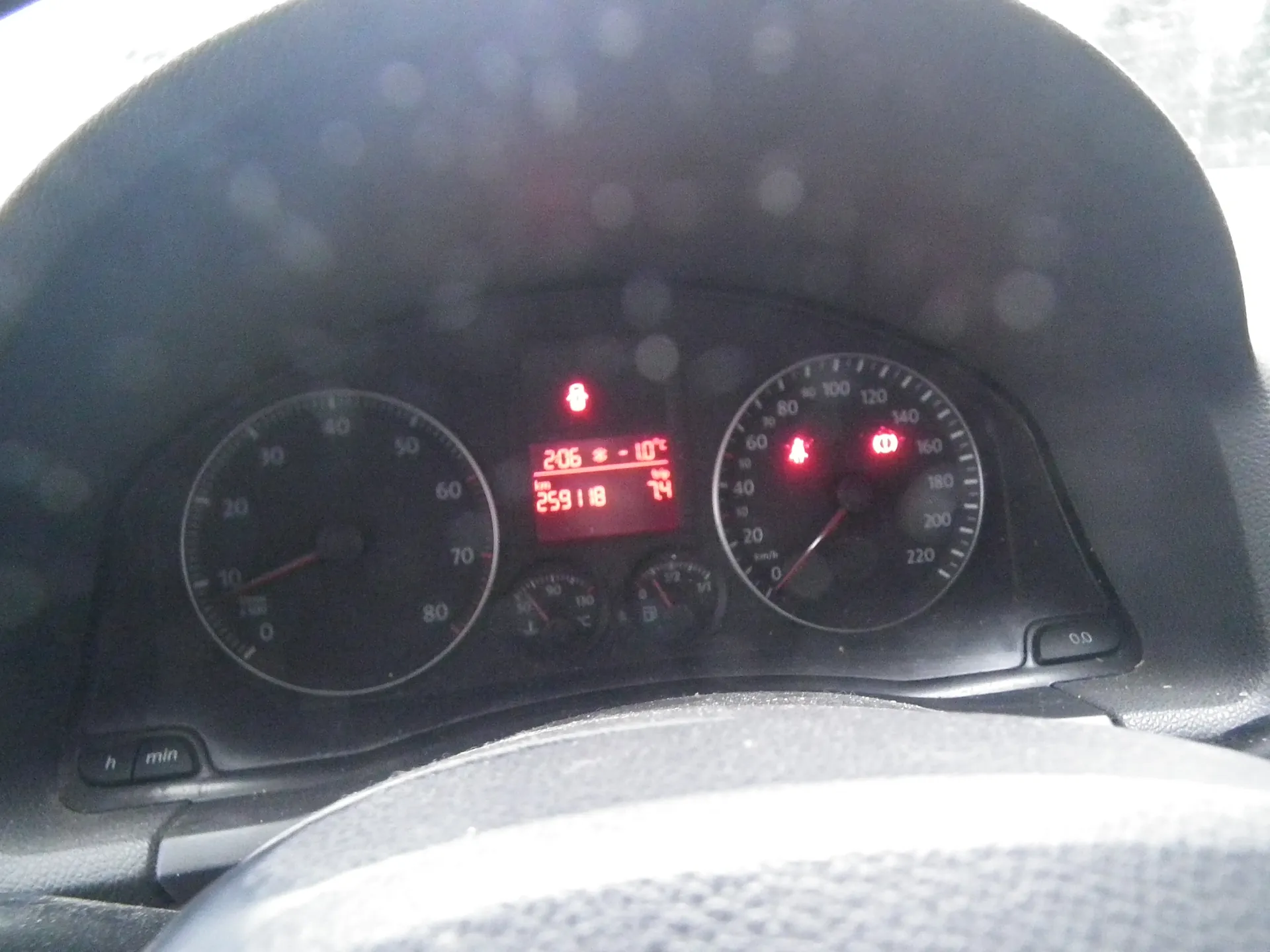 Tablica rozdzielcza samochodu z czarnym tłem. Po lewej stronie wskaźnik obrotomierza z zakresem 0-80, po prawej prędkościomierz z zakresem 0-220 km/h. W centrum czerwony wyświetlacz cyfrowy z danymi: 206, 259118, 74 oraz temperatura -10°C. Widoczne czerwone lampki ostrzegawcze, w tym ikona pasów bezpieczeństwa i innych symboli. Na dole fragment kierownicy. Po lewej stronie przyciski oznaczone 'h' i 'min'. Na wyświetlaczu i tle widoczne plamy światła.