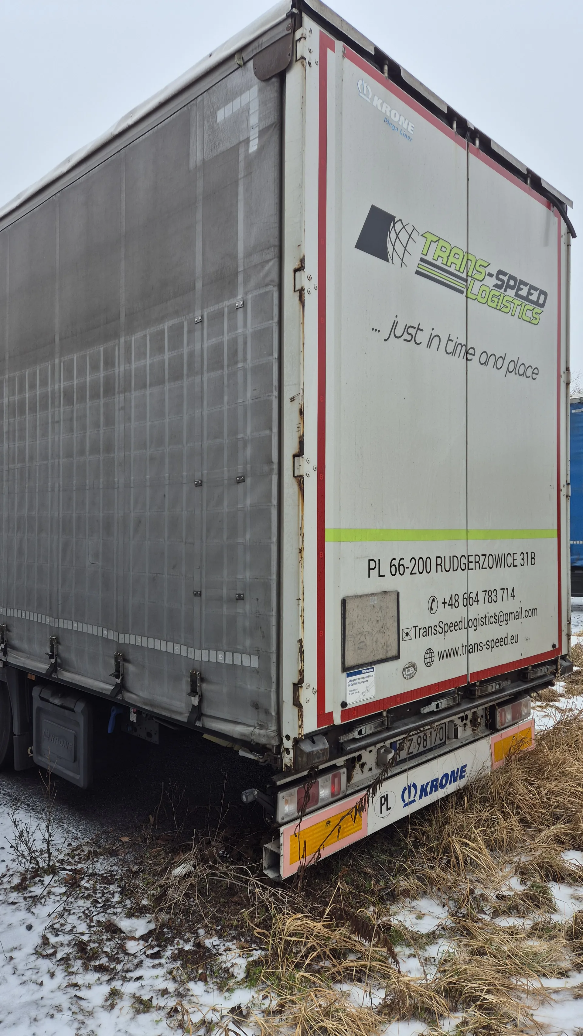 Biały naczepa ciężarowa z logo "TRANS-SPEED LOGISTICS" w kolorach zielonym i czarnym, hasłem "...just in time and place", adresem "PL 66-200 RUDGERZOWICE 31B", numerem telefonu "+48 664 783 714", adresem e-mail "TransSpeedLogistics@gmail.com" oraz stroną internetową "www.trans-speed.eu". Na boku naczepy widoczne logo "KRONE". Pojazd zaparkowany na śnieżnej powierzchni z suchą trawą, pod pochmurnym niebem.