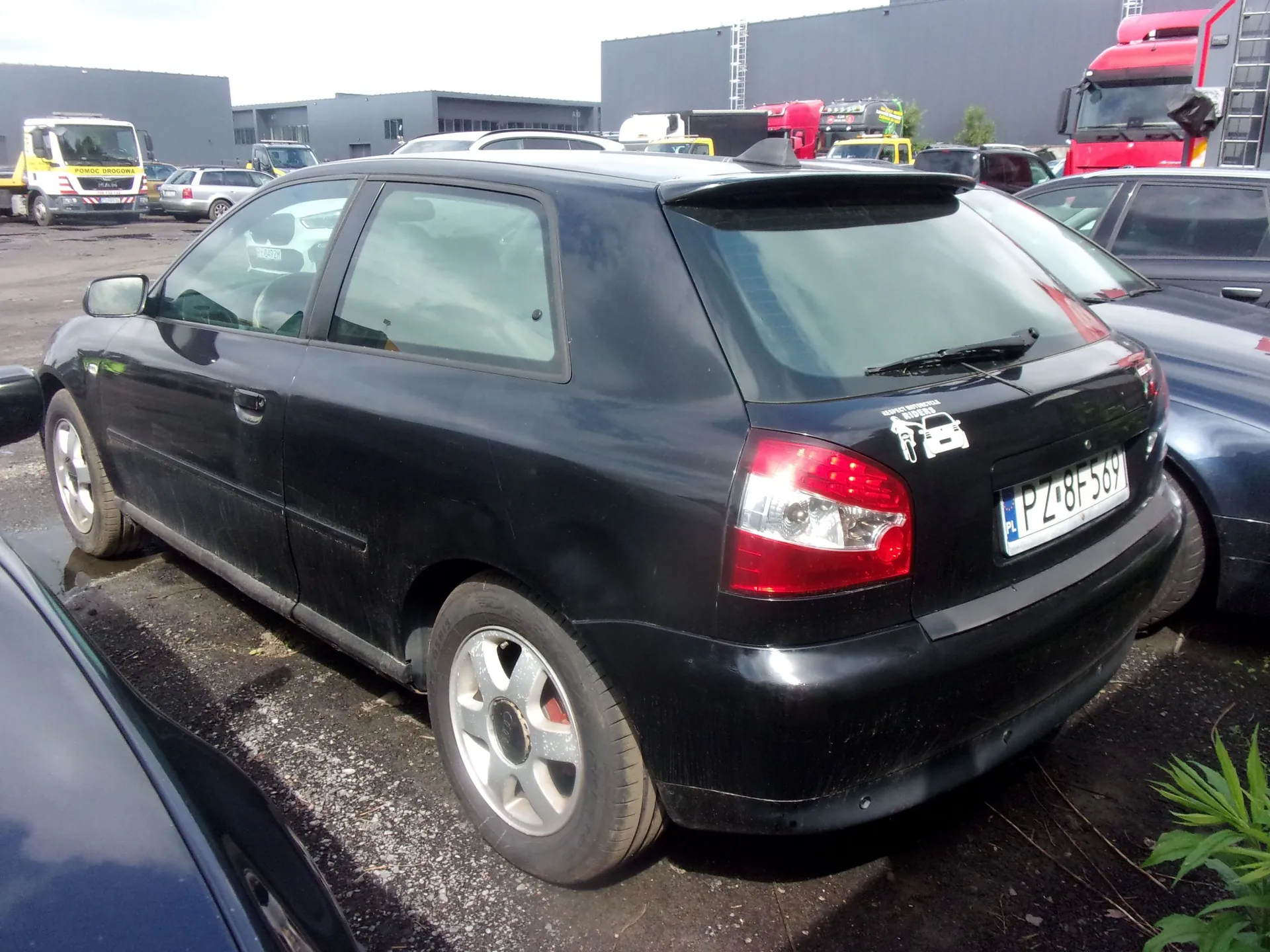 Czarny samochód osobowy typu hatchback zaparkowany na placu. Widoczny jest tył i bok pojazdu. Samochód ma polską tablicę rejestracyjną o numerze PZ 8BF569 oraz naklejkę z sylwetką samochodu na tylnej klapie. W tle widoczne są inne zaparkowane auta, w tym biały i czerwony samochód ciężarowy, oraz nowoczesny budynek przemysłowy.