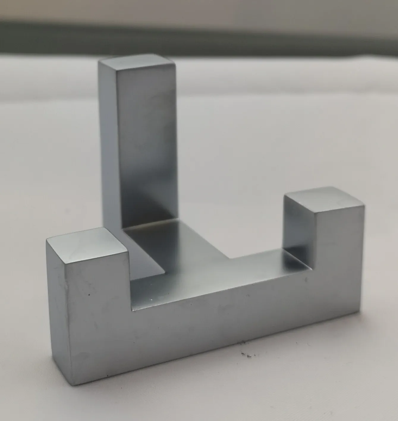 Srebrny, metalowy element konstrukcyjny o geometrycznym kształcie, przypominający literę U z dodatkowym pionowym słupkiem wyrastającym z wewnętrznej podstawy. Przedmiot znajduje się na białym tle.