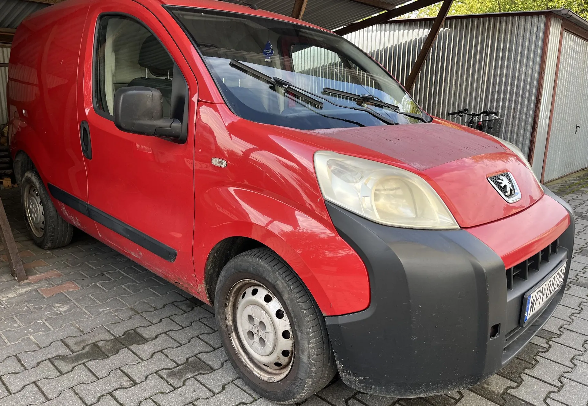Czerwony samochód dostawczy marki Peugeot zaparkowany na kostce brukowej pod wiatą. Widoczny jest przód i lewy bok pojazdu. Samochód ma czarny zderzak i czarne listwy boczne. W tle znajduje się metalowa ściana garażu.