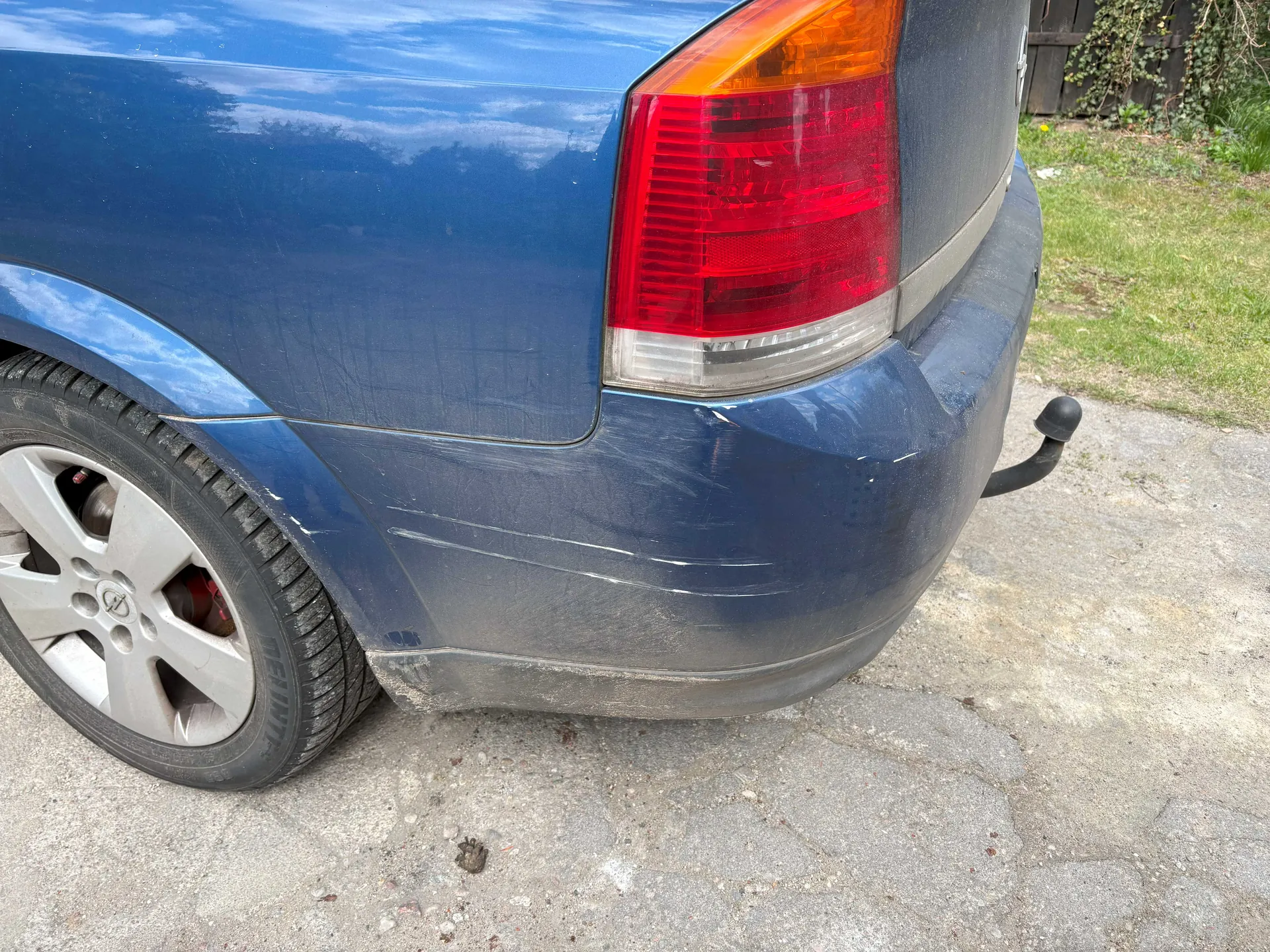 Zbliżenie na tylną część niebieskiego samochodu. Widoczny jest czerwony i pomarańczowy kierunkowskaz, zarysowane zadrapania na błotniku oraz brud na zderzaku. Koło z srebrną felgą i czarną oponą, na tle betonowej nawierzchni, trawy i drewnianego ogrodzenia z pnącza. Na karoserii odbijają się chmury i niebo.