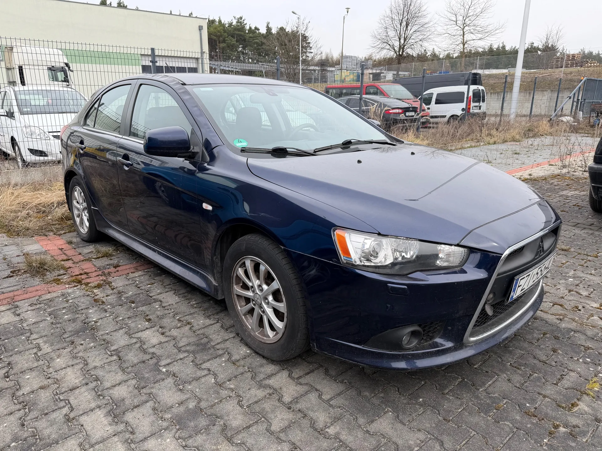 Ciemnoniebieski samochód osobowy marki Mitsubishi z tablicą rejestracyjną EZ 1557H, zaparkowany na brukowanym placu. Na przedniej szybie widoczny zielony naklejony znaczek. W tle ogrodzenie siatkowe, za którym stoją inne pojazdy: biały samochód, czerwony naczepowy pojazd, biały van oraz ciemny samochód. Po lewej stronie budynek z zielonym pasem, drzewa bez liści i słupy oświetleniowe. Podłoga z betonowych płyt z czerwonymi wstawkami, sucha trawa przy ogrodzeniu.