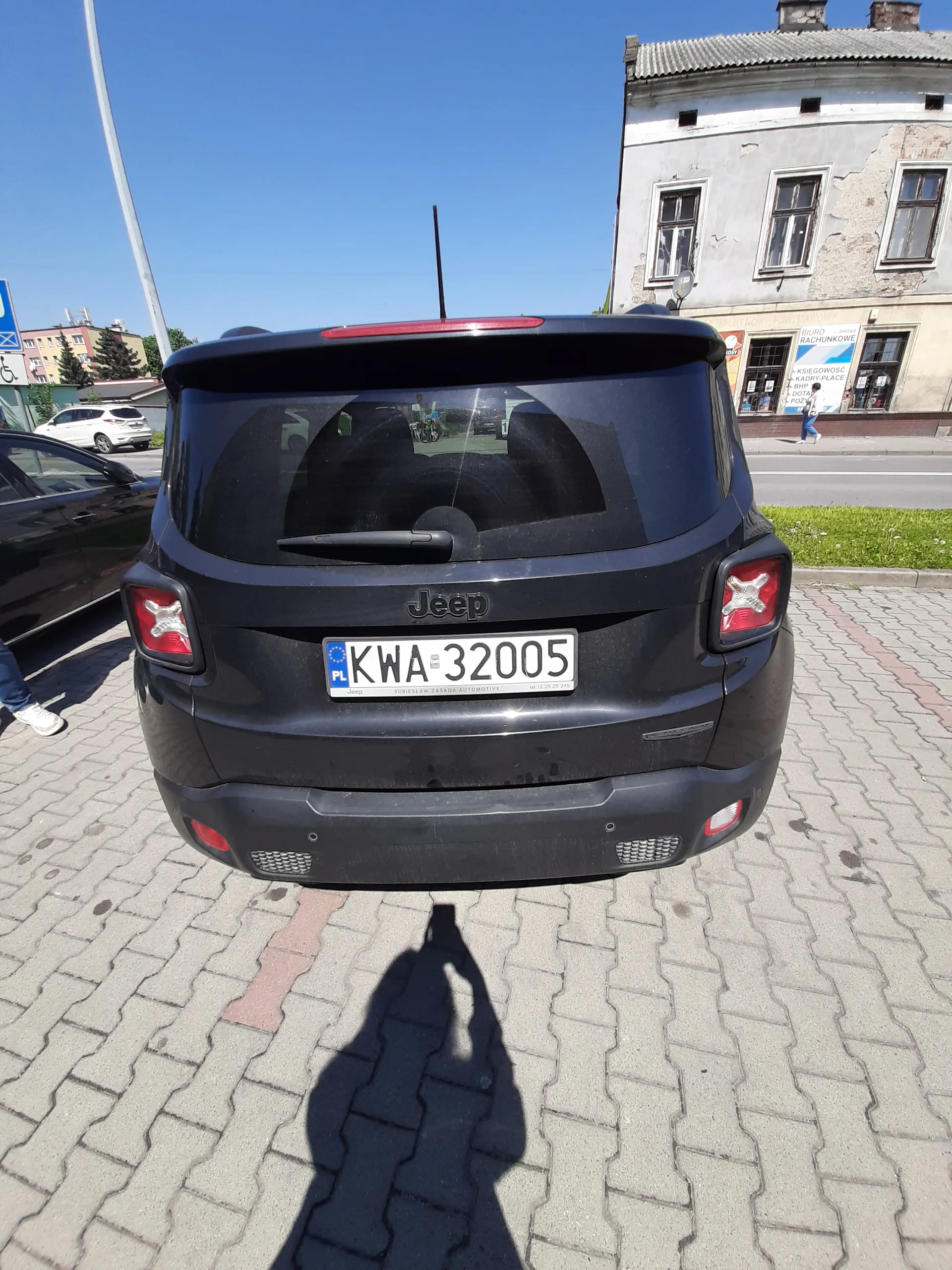 Tył ciemnoniebieskiego samochodu Jeep z tablicą rejestracyjną KWA 32005. Samochód zaparkowany na chodniku z kostki brukowej. W tle budynek z obłupioną farbą, oknami i niebieskim niebem. Widoczny cień osoby na chodniku oraz fragment innych pojazdów. Tablica rejestracyjna zawiera napis PL oraz numer KWA 32005.