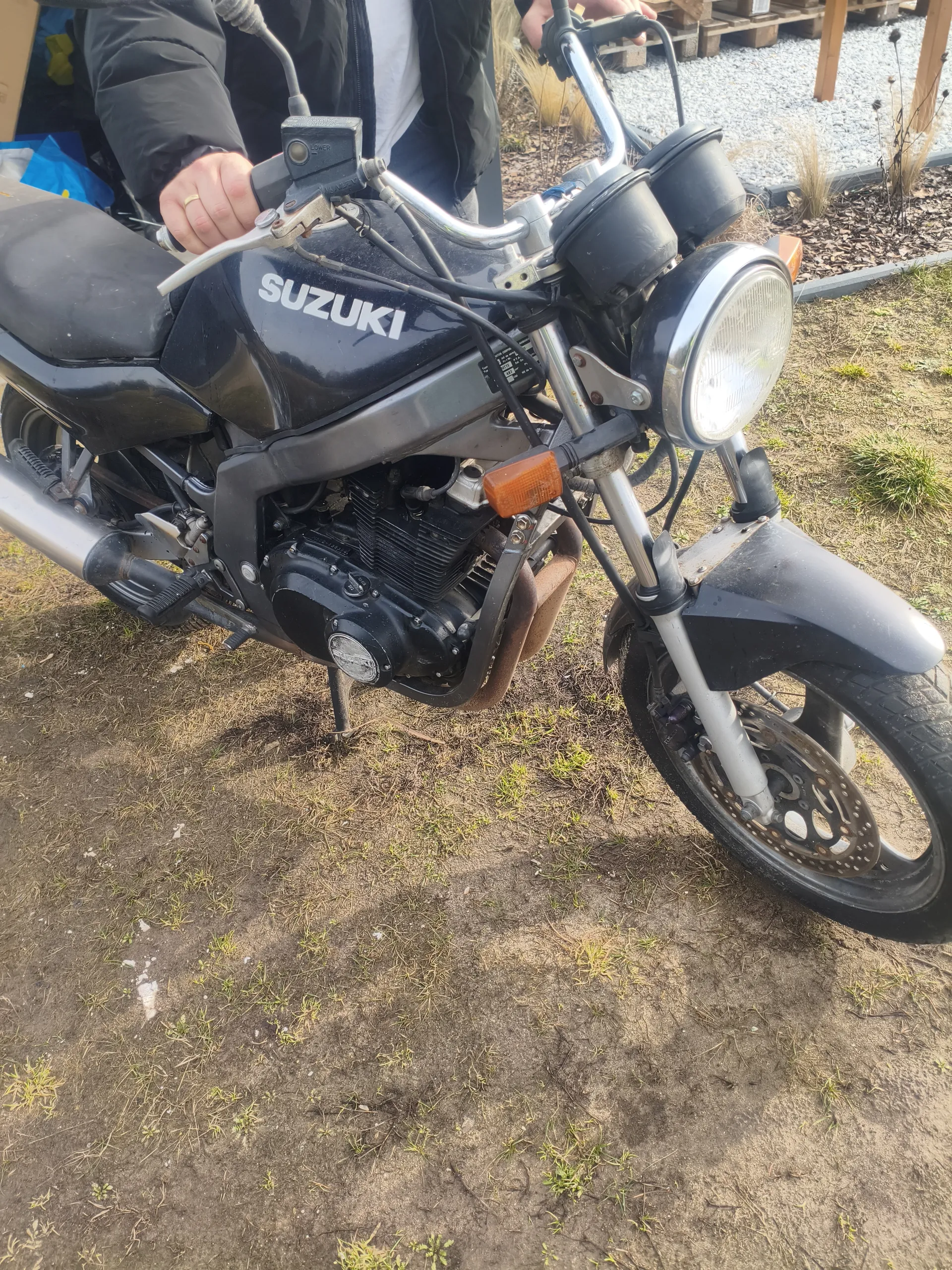 Czarny motocykl marki SUZUKI z widocznym silnikiem, reflektorem i kierownicą. Osoba w czarnej kurtce i białej koszulce trzyma kierownicę. Tło: sucha trawa, żwir i drewniane palety.