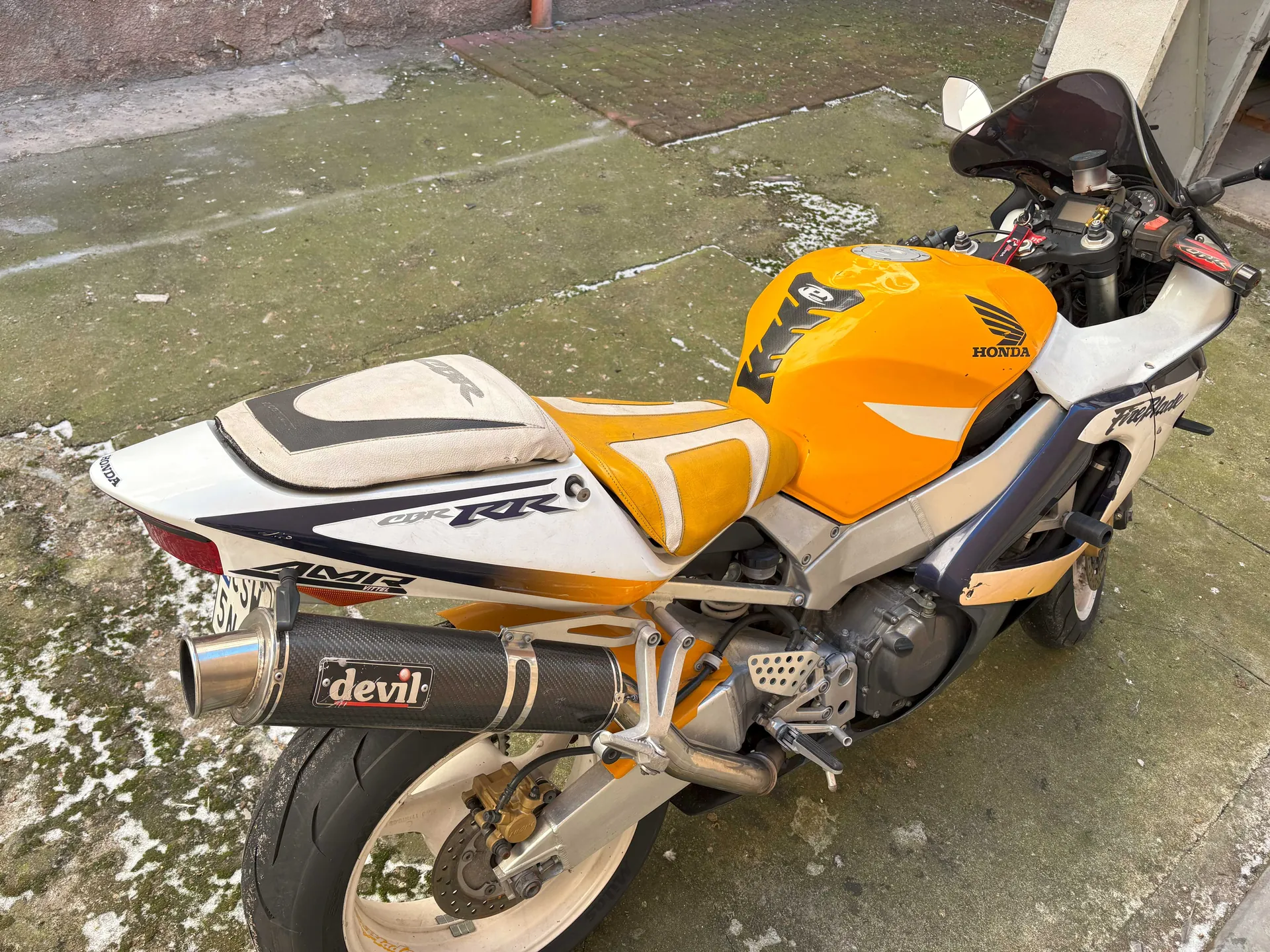 Motocykl marki Honda model CBR, kolor żółty z białymi i czarnymi elementami. Widoczne logo Honda na zbiorniku paliwa, napis Fireblade na boku, a na wydechu napis devil. Motocykl stoi na betonowej powierzchni z plamami mchu i śniegu. Widoczne są również tablica rejestracyjna, siedzenie z czarno-białym wzorem oraz elementy mechaniczne, w tym tylne koło z hamulcem tarczowym.