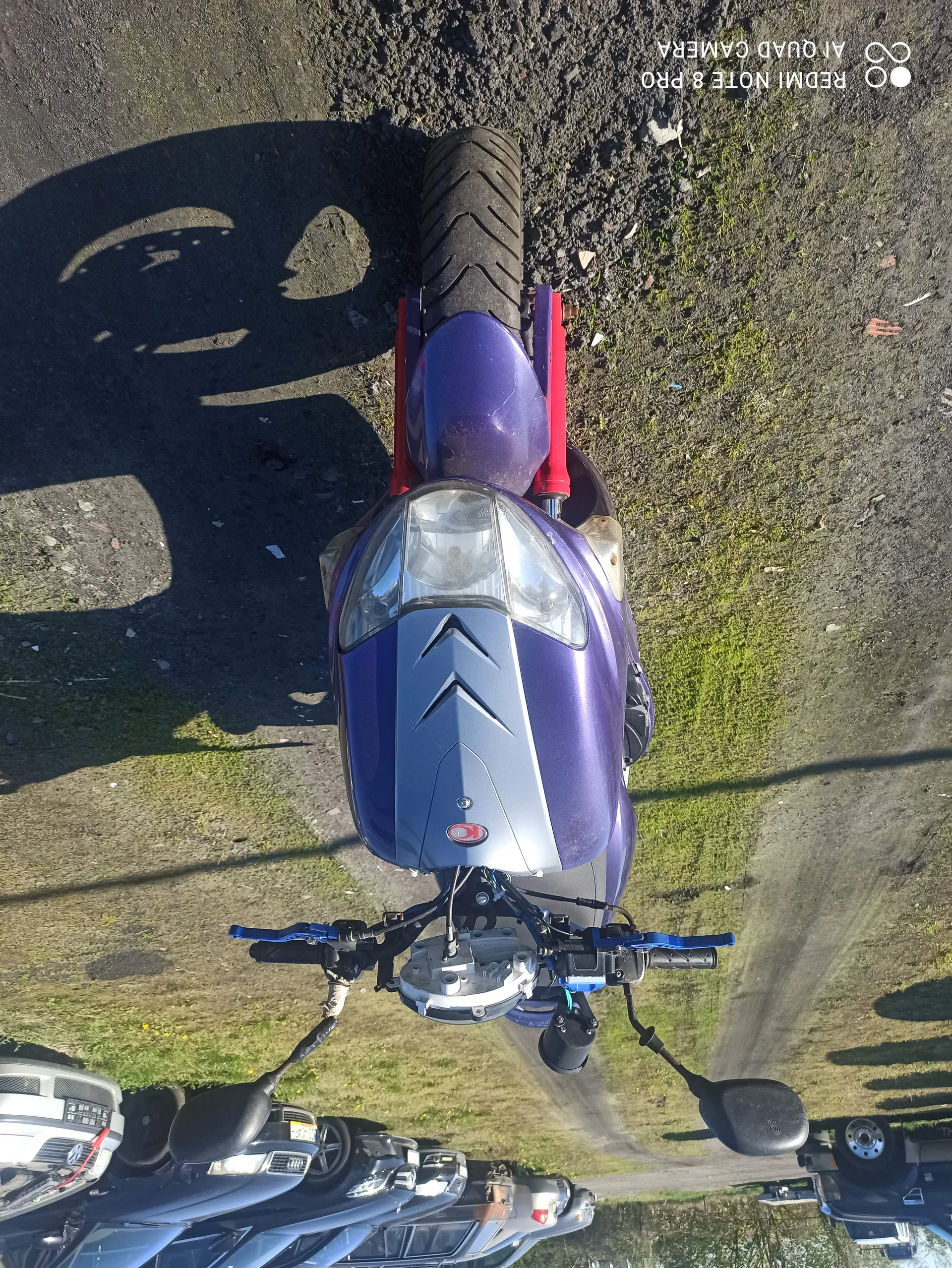 Fioletowy i srebrny skuter z czerwoną ramą, niebieskimi uchwytami kierownicy, reflektorem i logo na przodzie. W tle widoczne są inne pojazdy, w tym samochody. Podłoże z trawą i ziemią, cienie na powierzchni. Po prawej stronie obrazu widoczny tekst: REDMI NOTE 8 PRO AI QUAD CAMERA