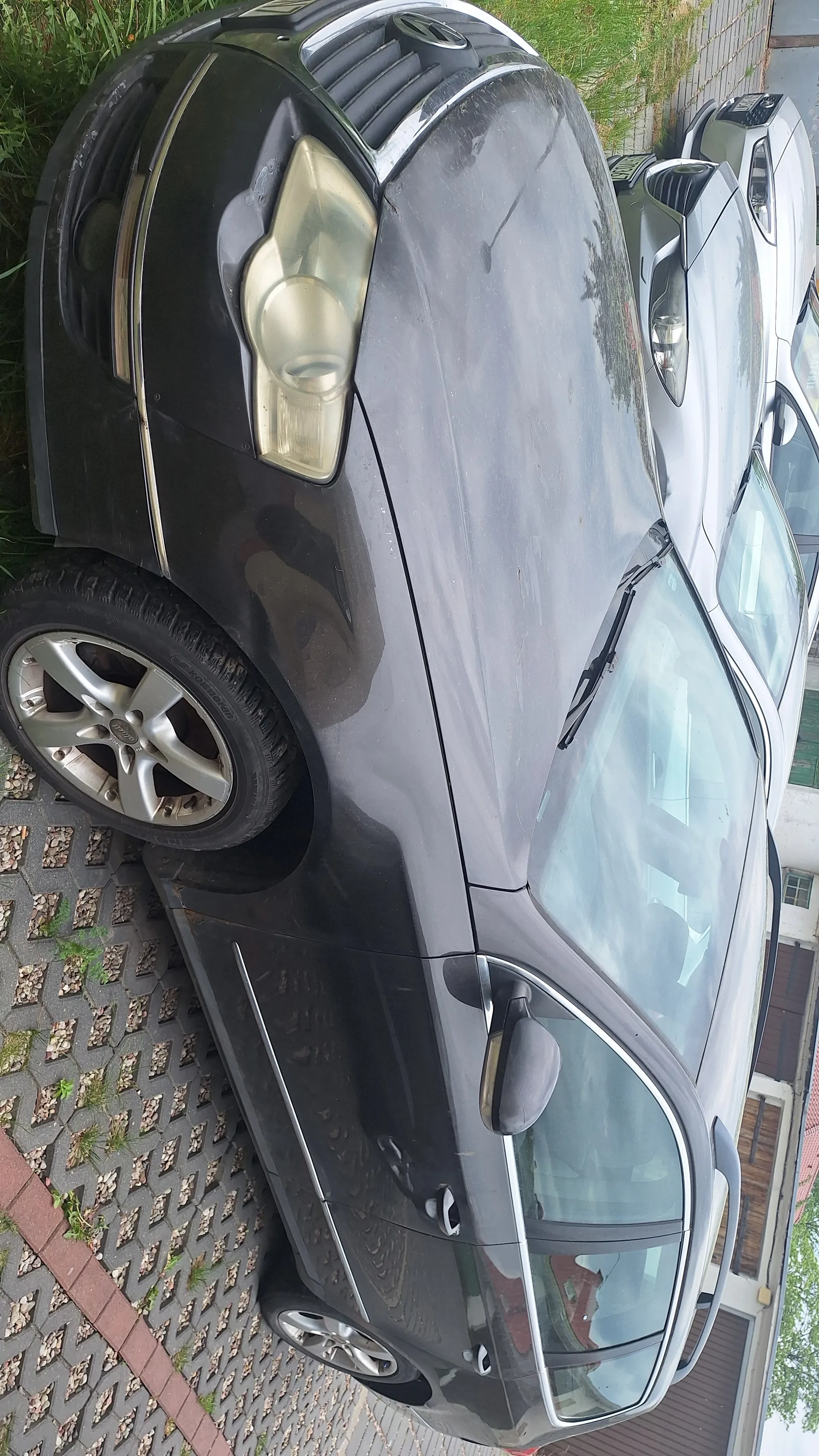 Czarny samochód osobowy zaparkowany na podwórku z kostki brukowej, widziany z boku. Widoczne są przednie światło, felgi aluminiowe oraz lusterko boczne. Obok czarnego samochodu znajduje się srebrny samochód z pionową tablicą rejestracyjną. W tle trawa i fragment budynku z brązową elewacją.