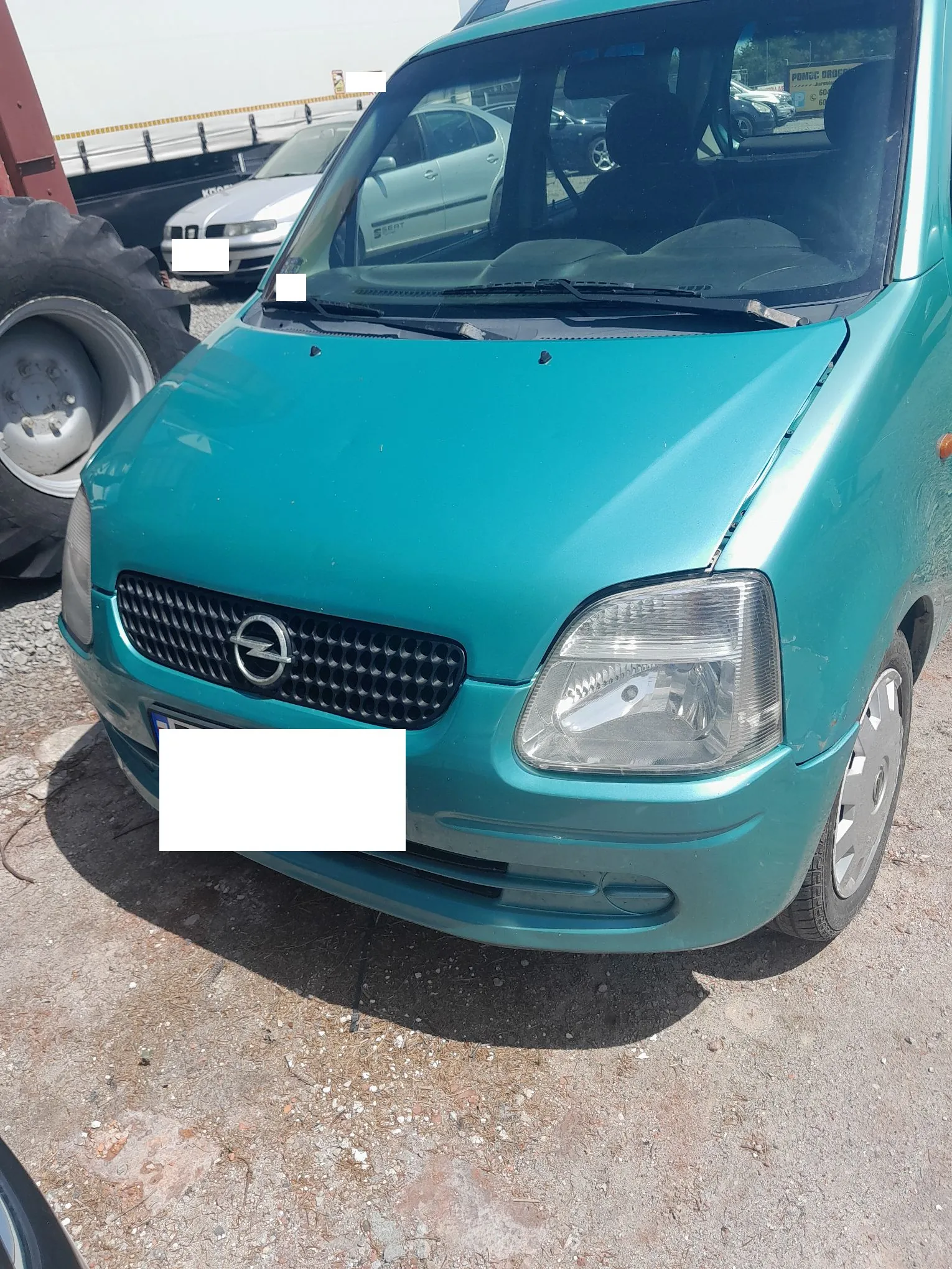 Zdjęcie przodu turkusowego samochodu marki Opel. Widoczny jest grill z logo producenta, reflektor po prawej stronie oraz maska z lekkim wgnieceniem. Tablica rejestracyjna jest zakryta białym prostokątem. W tle widoczne są inne pojazdy, w tym srebrny samochód oraz duże koło traktora po lewej stronie.