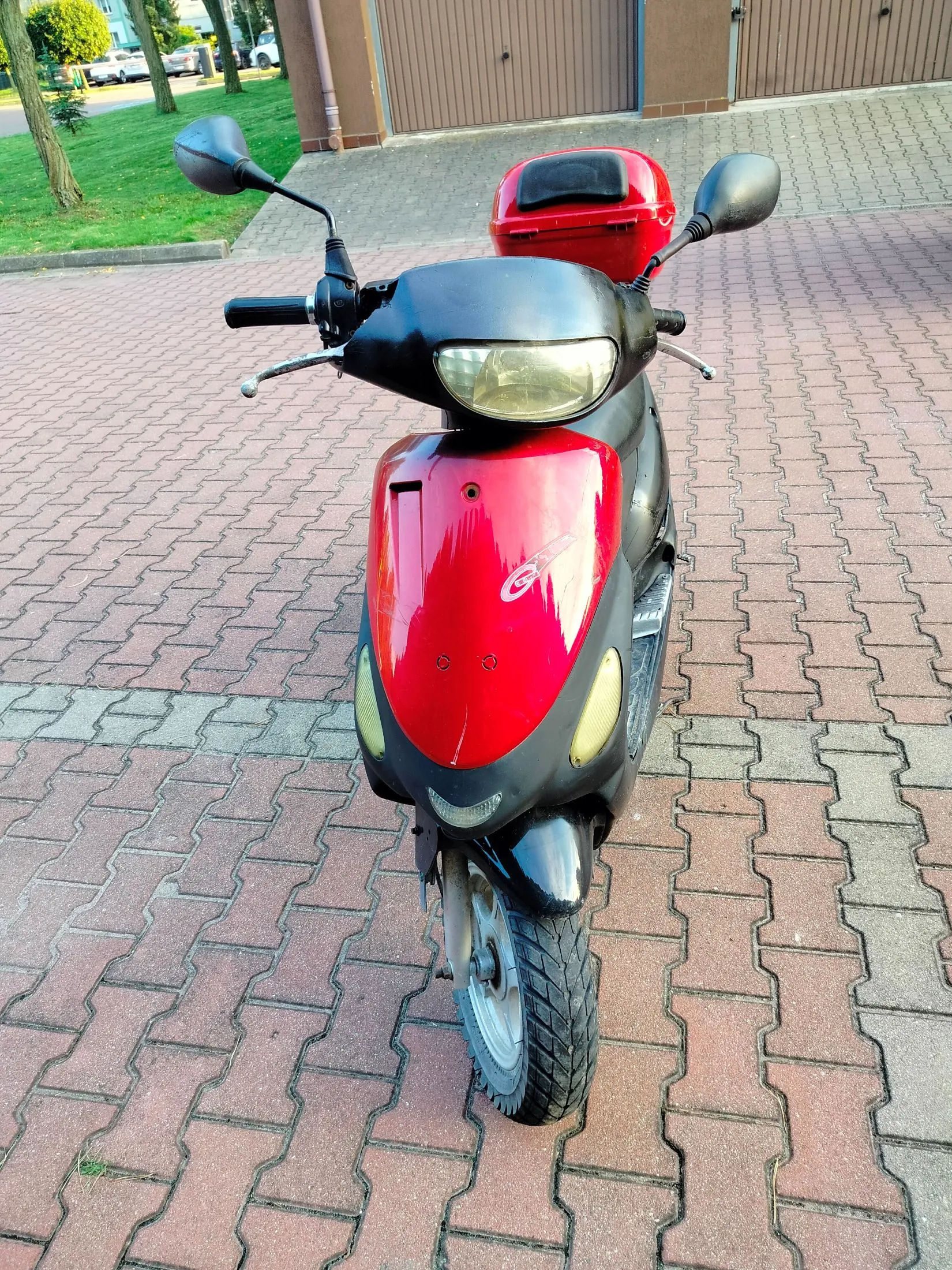 Czerwono-czarny skuter zaparkowany na kostce brukowej. Pojazd jest ustawiony przodem do widza, posiada przedni reflektor, lusterka boczne oraz czerwony kufer zamykany na klucz zamontowany z tyłu. W tle widoczny jest fragment budynku z bramami garażowymi oraz trawnik z drzewami.