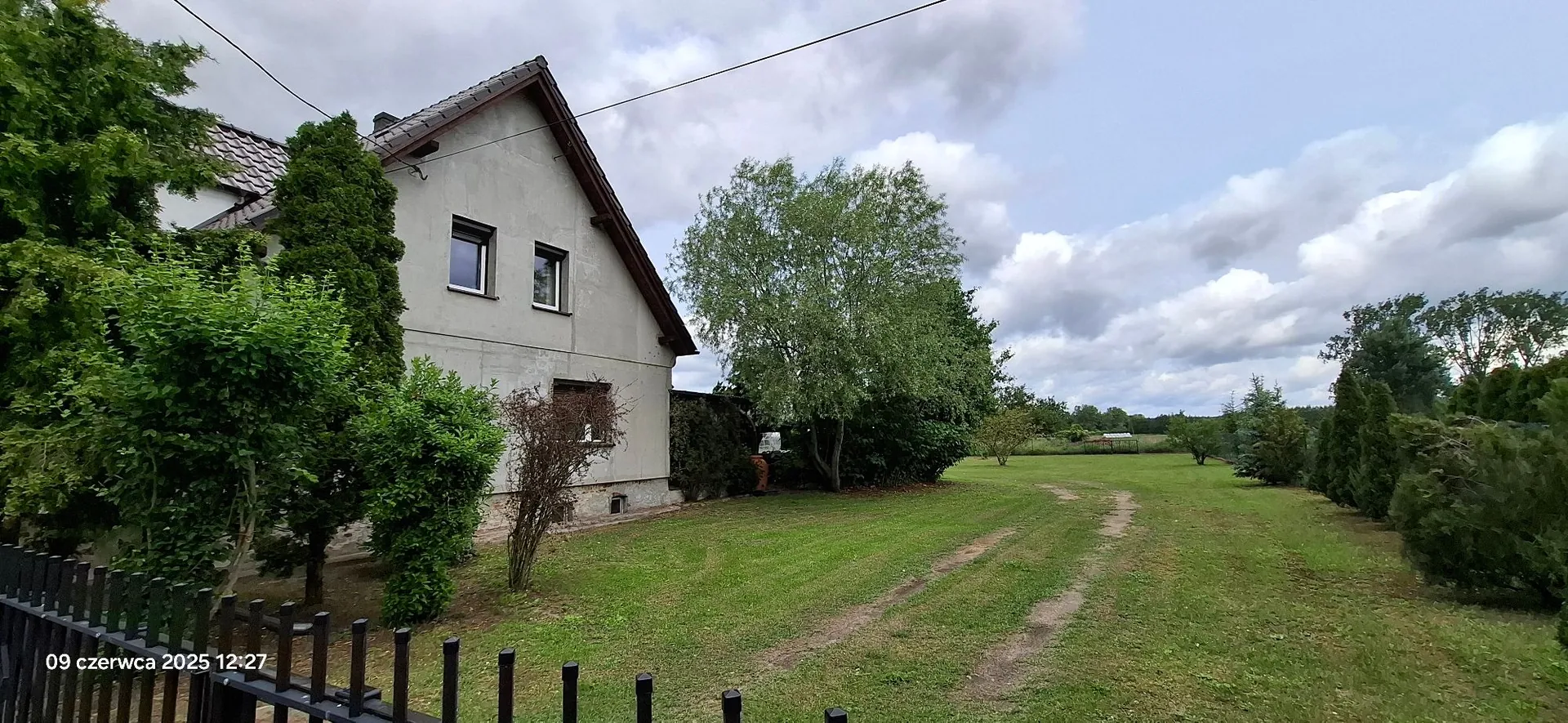 Zdjęcie przedstawia jednokondygnacyjny dom z poddaszem o szarych ścianach i spadzistym dachem. Obok domu rosną wysokie, zielone drzewa i krzewy. Przed budynkiem znajduje się rozległy, zielony trawnik z widocznymi śladami kół samochodu prowadzącymi w głąb posesji. W lewym dolnym rogu widoczny jest fragment czarnego metalowego ogrodzenia oraz biały napis z datą i godziną: '09 czerwca 2025 12:27'. Niebo jest zachmurzone.