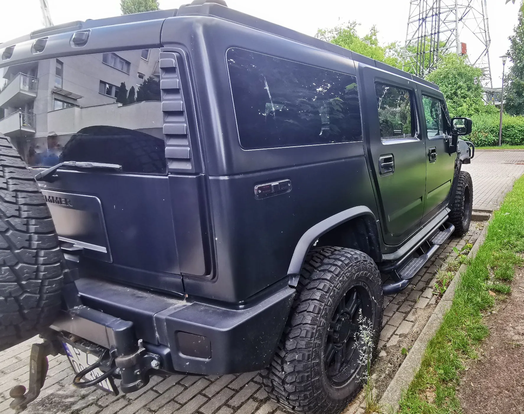 Czarny, matowy samochód terenowy marki Hummer zaparkowany na kostce brukowej obok trawnika. Widoczny jest tył i prawy bok pojazdu, w tym zapasowe koło zamontowane z tyłu, szerokie opony terenowe oraz stopnie boczne. W odbiciu szyb widoczne są budynki mieszkalne.