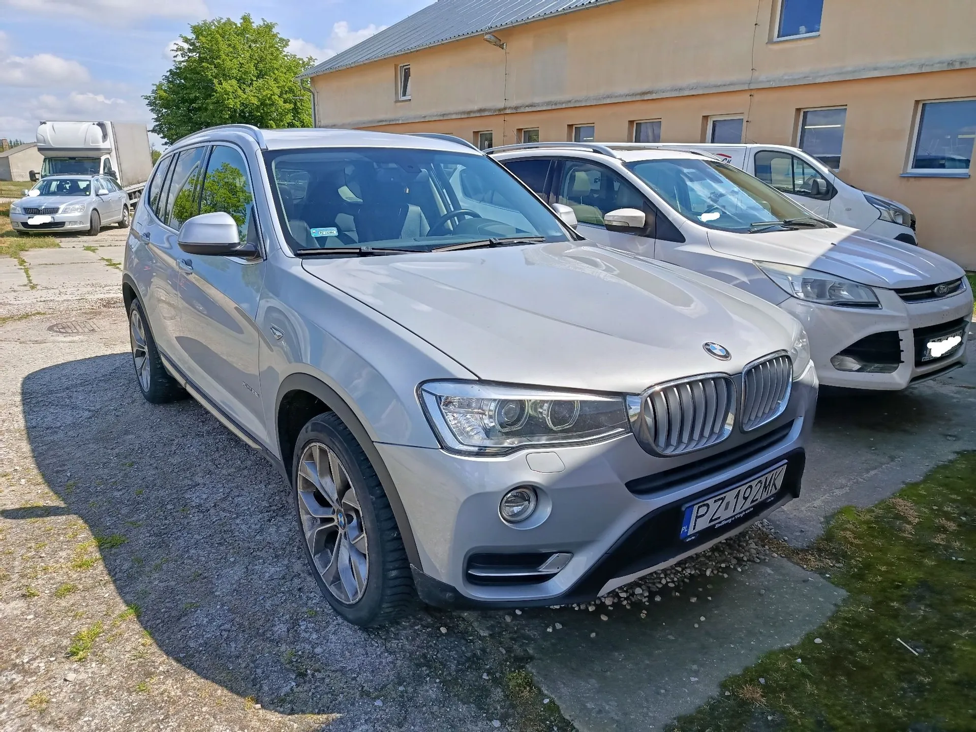 Srebrny samochód marki BMW X5 zaparkowany na betonowym placu. Obok niego stoi biały samochód marki Ford. W tle widoczny jest żółty budynek oraz biała ciężarówka. Samochód BMW ma polską tablicę rejestracyjną o numerze PZ 192MK.