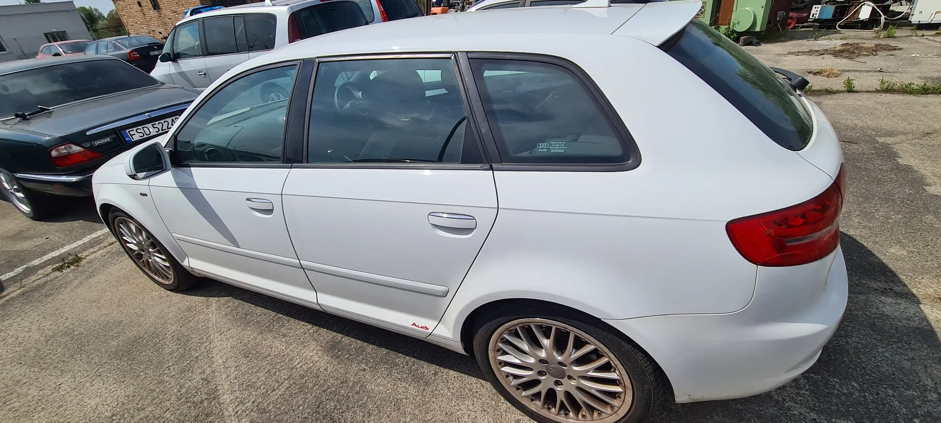 Biały samochód Audi typu hatchback z pięcioma drzwiami, z srebrnymi felgami i czerwonym napisem "Audi" na boku. Tylne szyby przyciemnione. W tle czarny samochód Jaguar z tablicą rejestracyjną FSD 5224, inne zaparkowane pojazdy, ceglany budynek oraz elementy przemysłowe na asfaltowym parkingu. Na tle widoczne są również fragmenty innych budynków i sucha trawa.