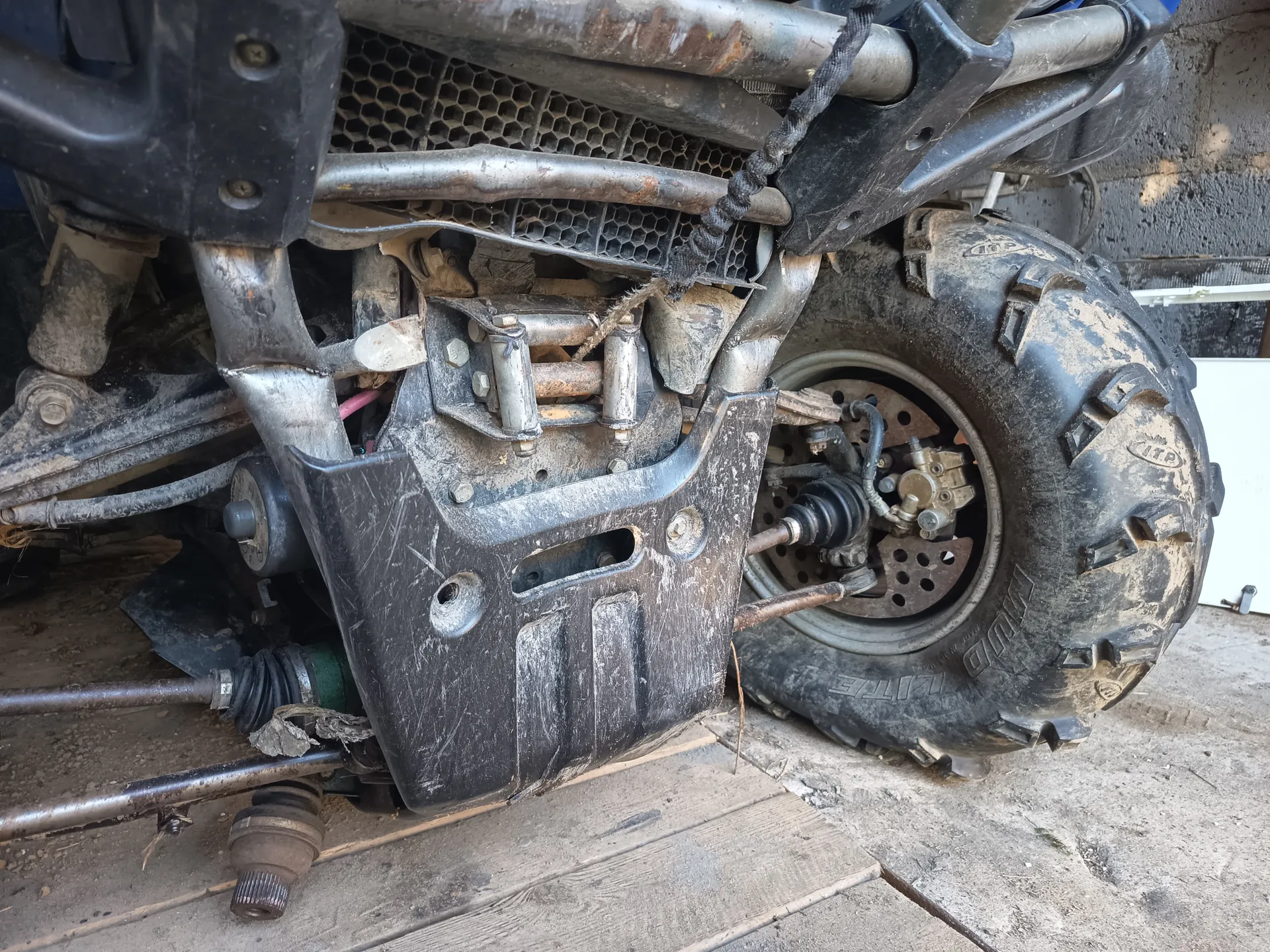 Zbliżenie na przednią część pojazdu terenowego, prawdopodobnie quadra. Widoczne są brudne metalowe elementy konstrukcyjne, miedziane rury, siatkowa osłona chłodnicy oraz duże opony z głębokim profilem. Na oponie widoczny napis 'ITP' oraz 'MUD LITE'. Pojazd znajduje się na betonowej powierzchni z drewnianymi deskami. Elementy są pokryte błotem i kurzem, a w tle widoczna jest betonowa ściana.
