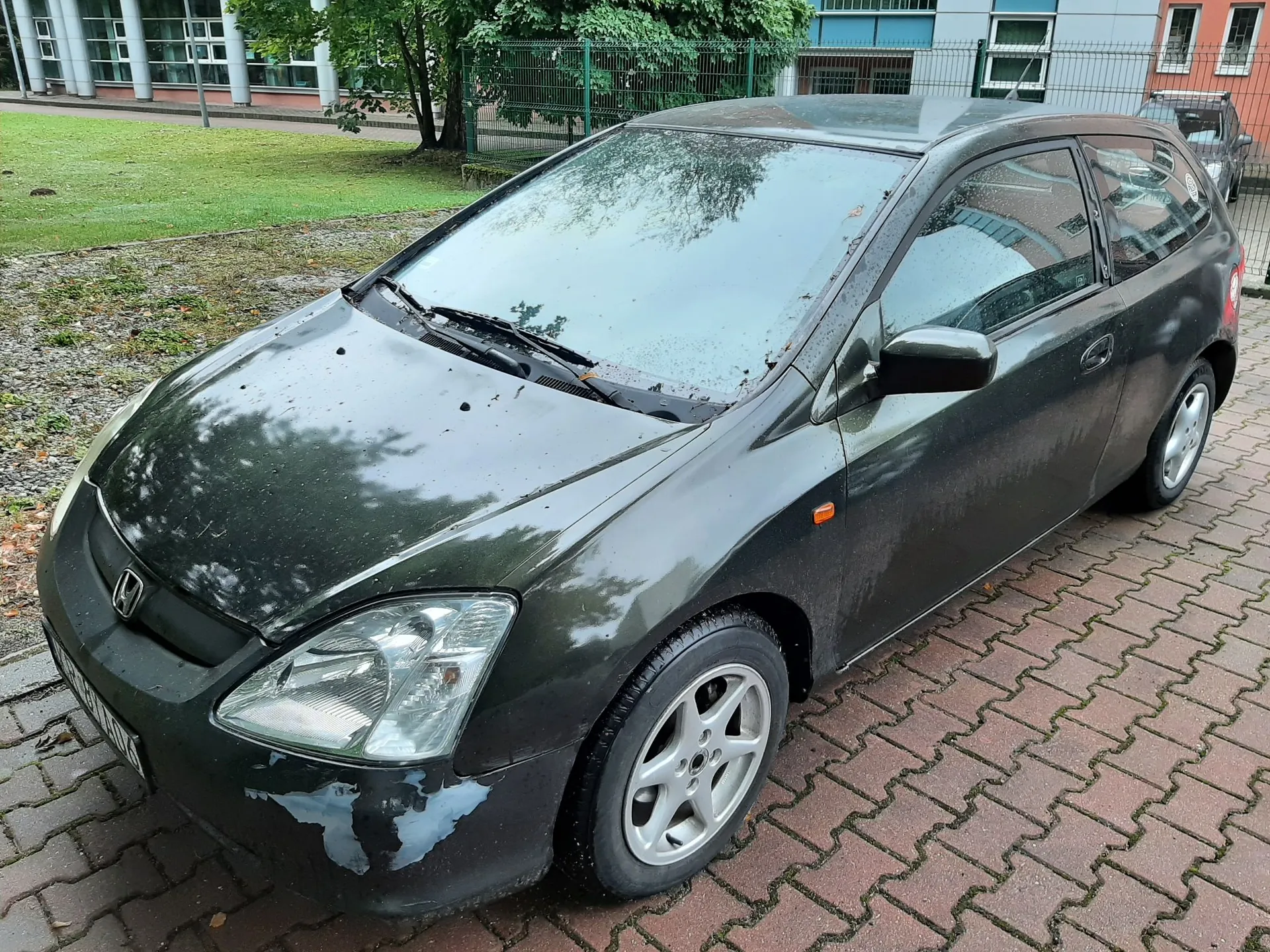 Ciemnozielony samochód osobowy marki Honda zaparkowany na kostce brukowej. Pojazd posiada widoczne uszkodzenia lakieru na przednim zderzaku po stronie kierowcy. W tle widać zielony trawnik, drzewa oraz fragmenty budynków.