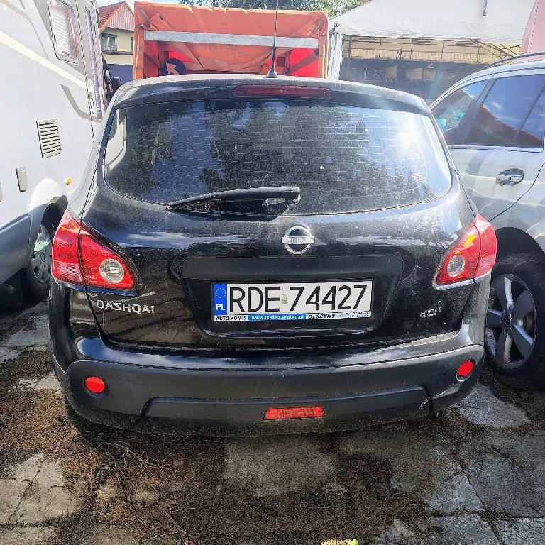 Tylny widok czarnego samochodu marki Nissan Qashqai. Pojazd posiada polską tablicę rejestracyjną o numerze RDE 74427. Po lewej stronie widoczny jest fragment białego kampera, a po prawej srebrny samochód. Auto stoi na utwardzonym podłożu z widocznymi resztkami roślinności.