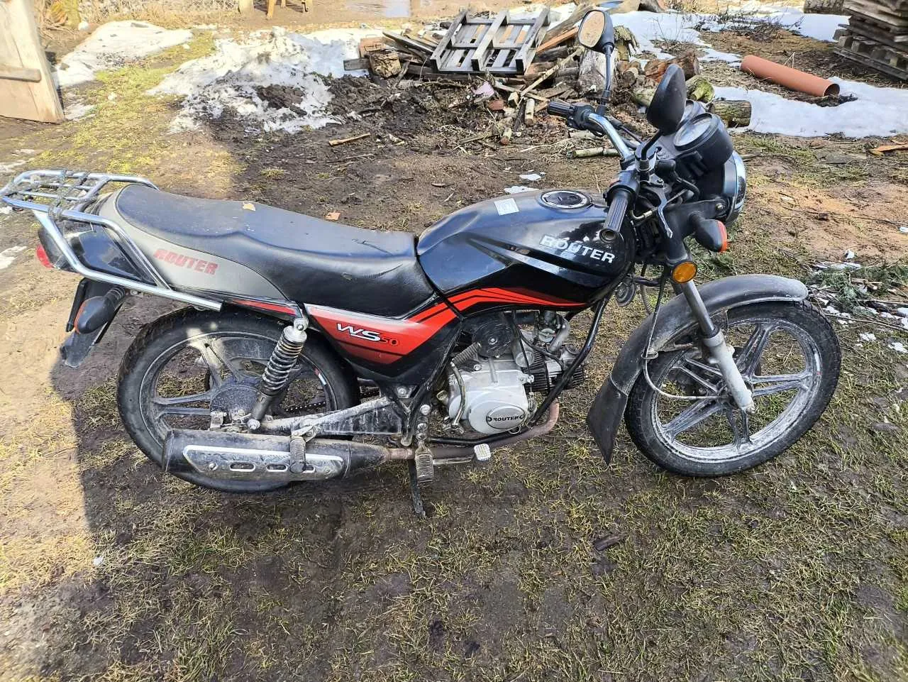 Czarny motocykl marki Rouser z czerwonymi akcentami, model WS, zaparkowany na trawiastym i błotnistym terenie. Na zbiorniku widoczny napis 'ROUTER', na boku 'WS'. W tle fragmenty śniegu, stosy drewna i palet. Teren otoczony suchą trawą i śniegiem.