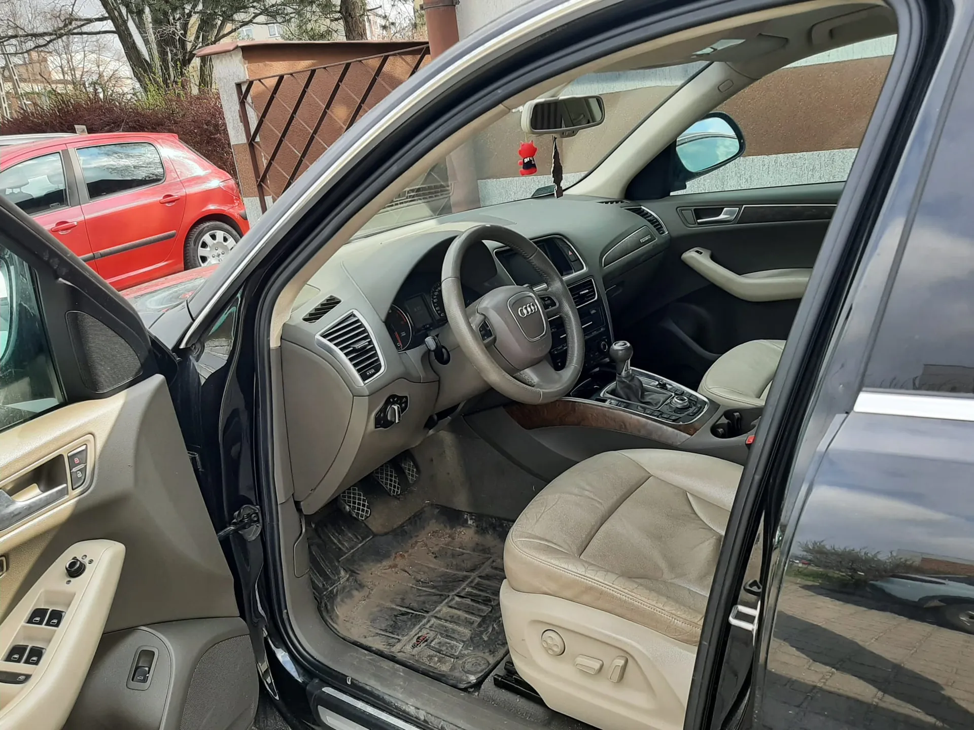 Wnętrze czarnego samochodu marki Audi z otwartą przodem drzwiami kierowcy. Widoczne są beżowe skórzane fotele, kierownica z logo Audi, deska rozdzielcza, dźwignia zmiany biegów oraz zabrudzone dywaniki. Na zewnątrz, w tle, stoi czerwony samochód, a za nim drzewa i budynki z ogrodzeniem. W lusterku wstecznym widoczny czerwony wiszący przedmiot.