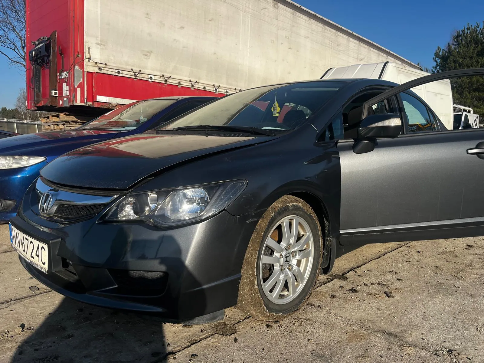 Ciemnoszary samochód marki Honda z otwartą przednią prawą drzwiami, z widoczną tablicą rejestracyjną HN 6724C. W tle niebieski samochód, czerwono-biały naczepa ciężarowa oraz betonowa powierzchnia z niebieskim niebem. Opona przedniego koła prawego jest zabrudzona błotem.