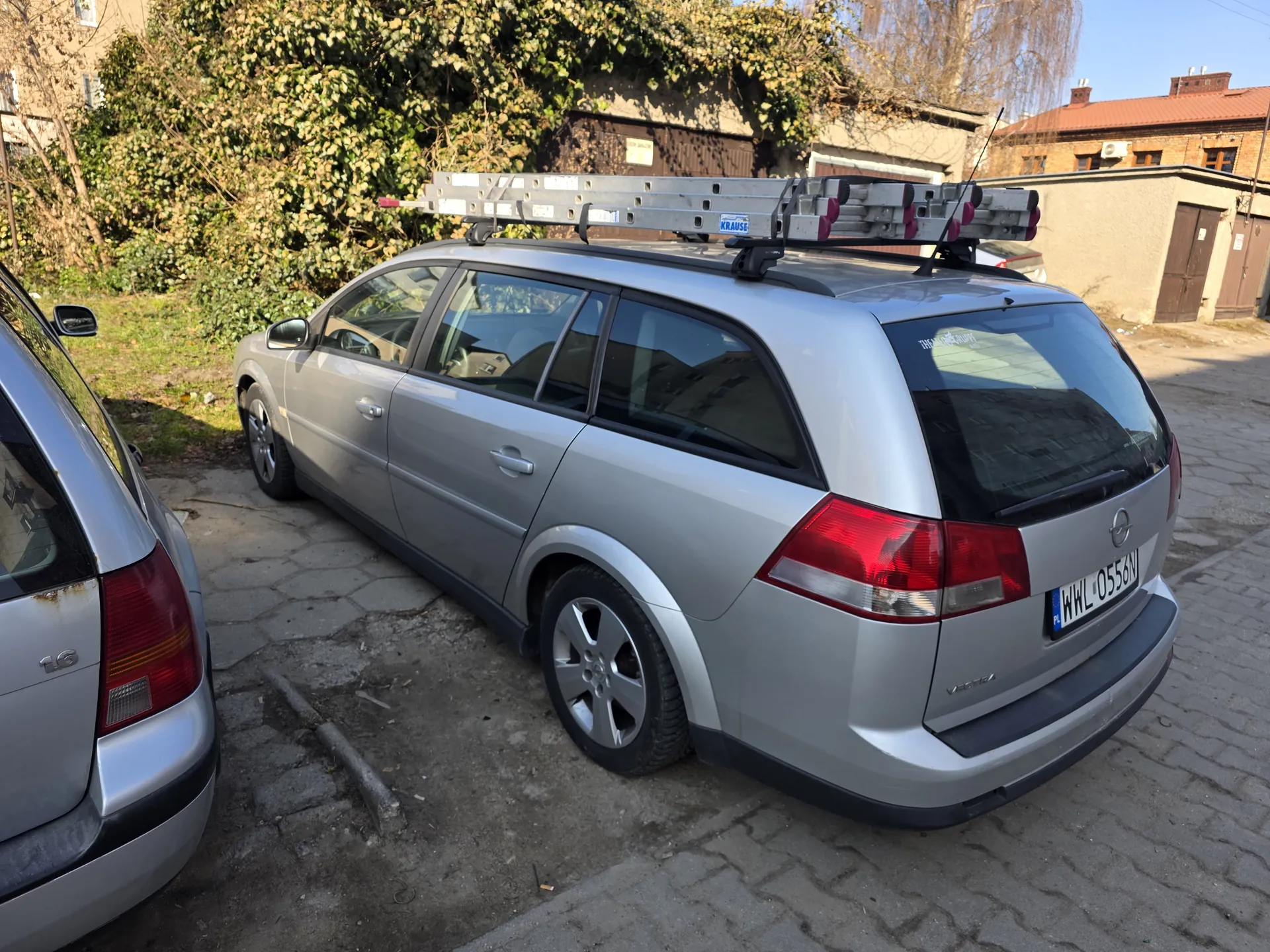 Srebrny samochód typu kombi marki Opel z dachowym uchwytem na drabiny. Widoczny tylny numer rejestracyjny WWL 0556M. Po lewej stronie częściowo widoczny inny srebrny samochód. Tło: zarośla, budynki mieszkalne z czerwonym dachem, brukowana powierzchnia. Na dachu głównego pojazdu umocowane są metalowe drabiny z widocznym napisem KRAUSE.