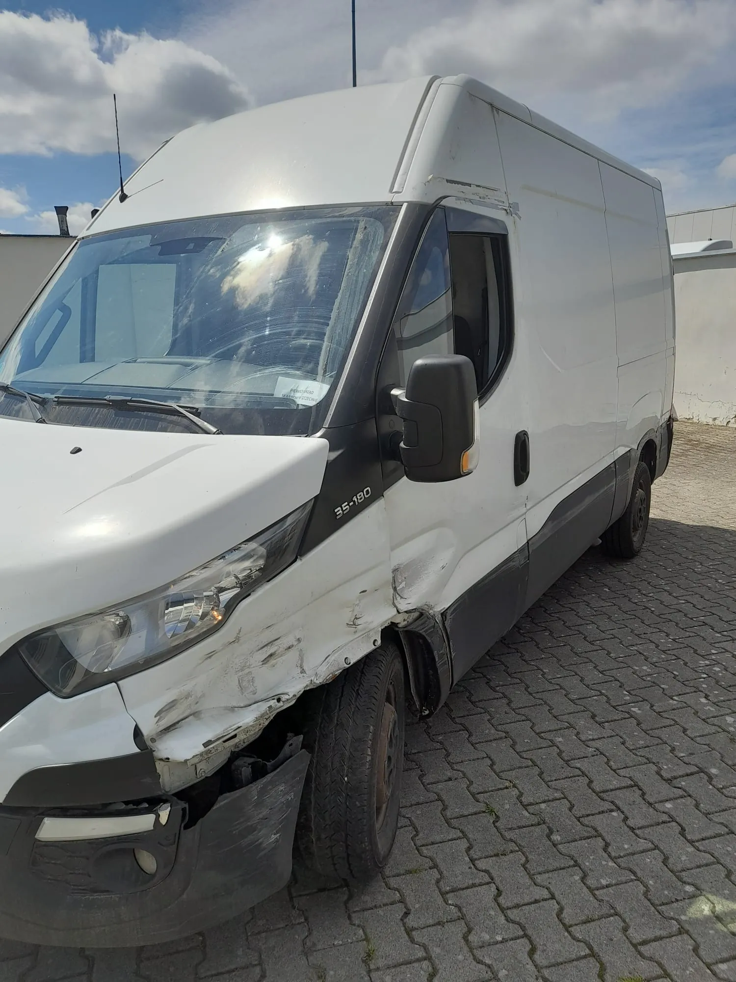 Biały furgon marki Iveco Daily z widocznymi uszkodzeniami karoserii po prawej stronie przodu. Zniszczony jest przedni zderzak oraz prawy błotnik, który jest wgnieciony. Na boku pojazdu widnieje napis '35-180'. Samochód stoi na kostce brukowej pod zachmurzonym niebem.