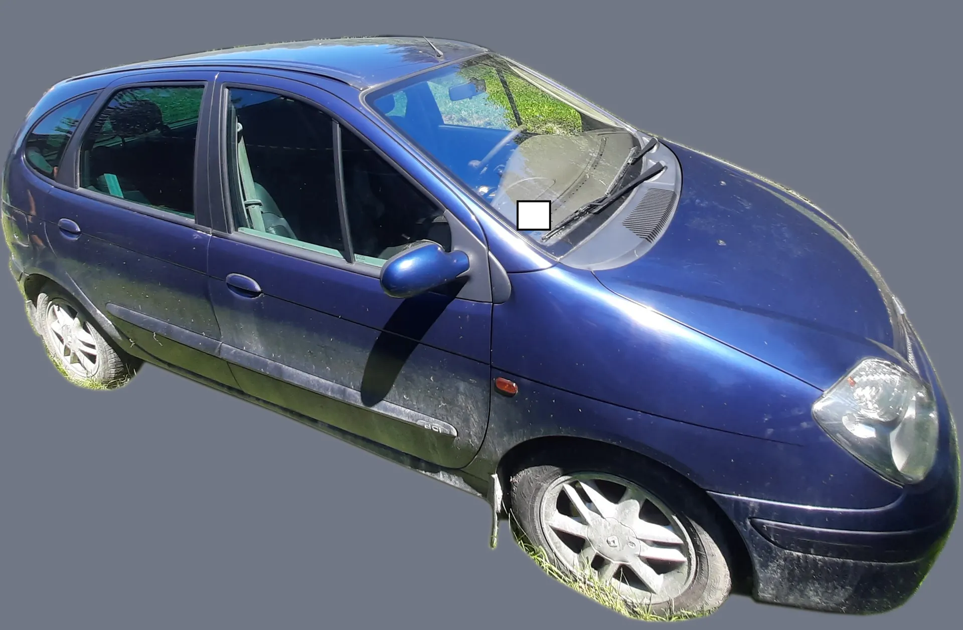 Niebieski samochód osobowy typu hatchback zaparkowany na trawie, widoczny z perspektywy przedniego lewego boku. Pojazd jest zakurzony, a na przedniej szybie znajduje się mały biały kwadratowy element.