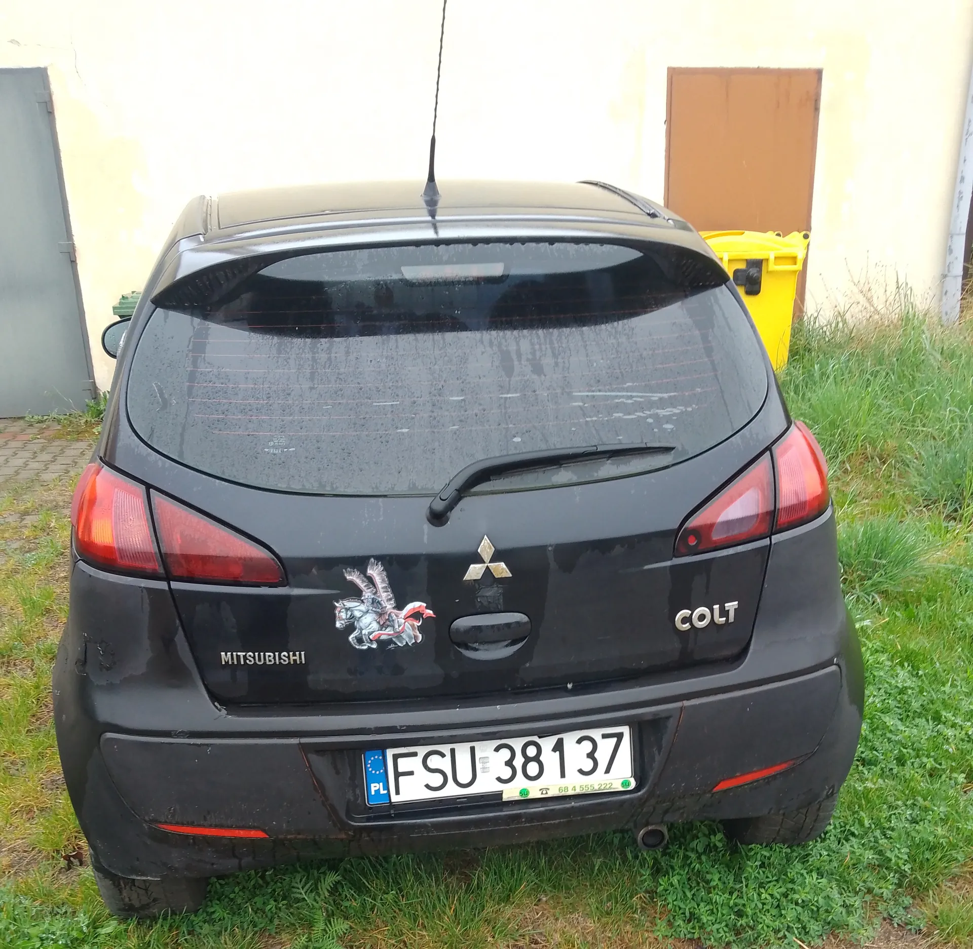 Zdjęcie przedstawia tył czarnego samochodu Mitsubishi Colt zaparkowanego na trawie. Na bagażniku widoczne są napisy "MITSUBISHI" po lewej stronie, logo trzech rombów w środku oraz napis "COLT" po prawej stronie. Poniżej znajduje się biała tablica rejestracyjna z polskim kodem "PL" i numerem "FSU 38137". Na lewej stronie bagażnika umieszczono naklejkę przedstawiającą jeźdźca na koniu. W tle widać żółty pojemnik na śmieci, brązowe drzwi oraz fragment budynku z jasną ścianą. Samochód ma wilgotną karoserię z widocznymi kroplami wody.
